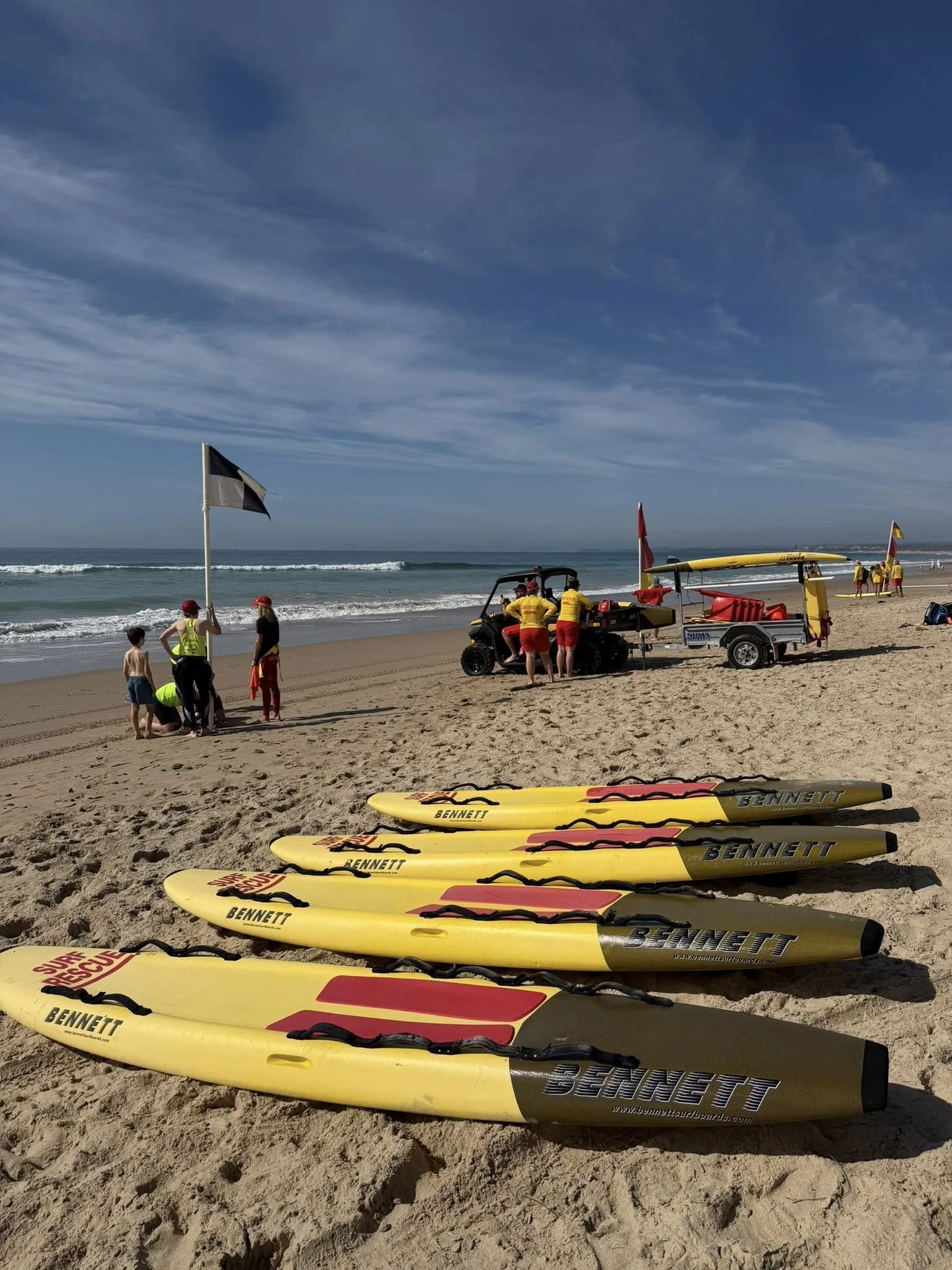 Yellow rescue surfboards lined up on the sandy beach, with people and rescue equipment in the background, under a partly cloudy sky.