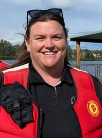 Woman smiling outdoors, wearing sunglasses on her head and a red and black uniform with a logo, standing near a body of water with a dock in the background.
