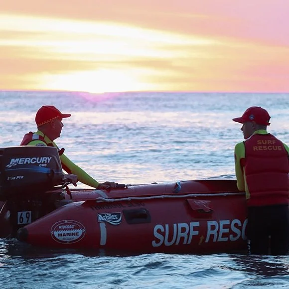 Two surf rescue personnel in wetsuits and red caps working on a red rescue boat in the water during sunset.