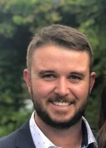 A man with short brown hair and a beard, smiling outdoors with trees in the background.