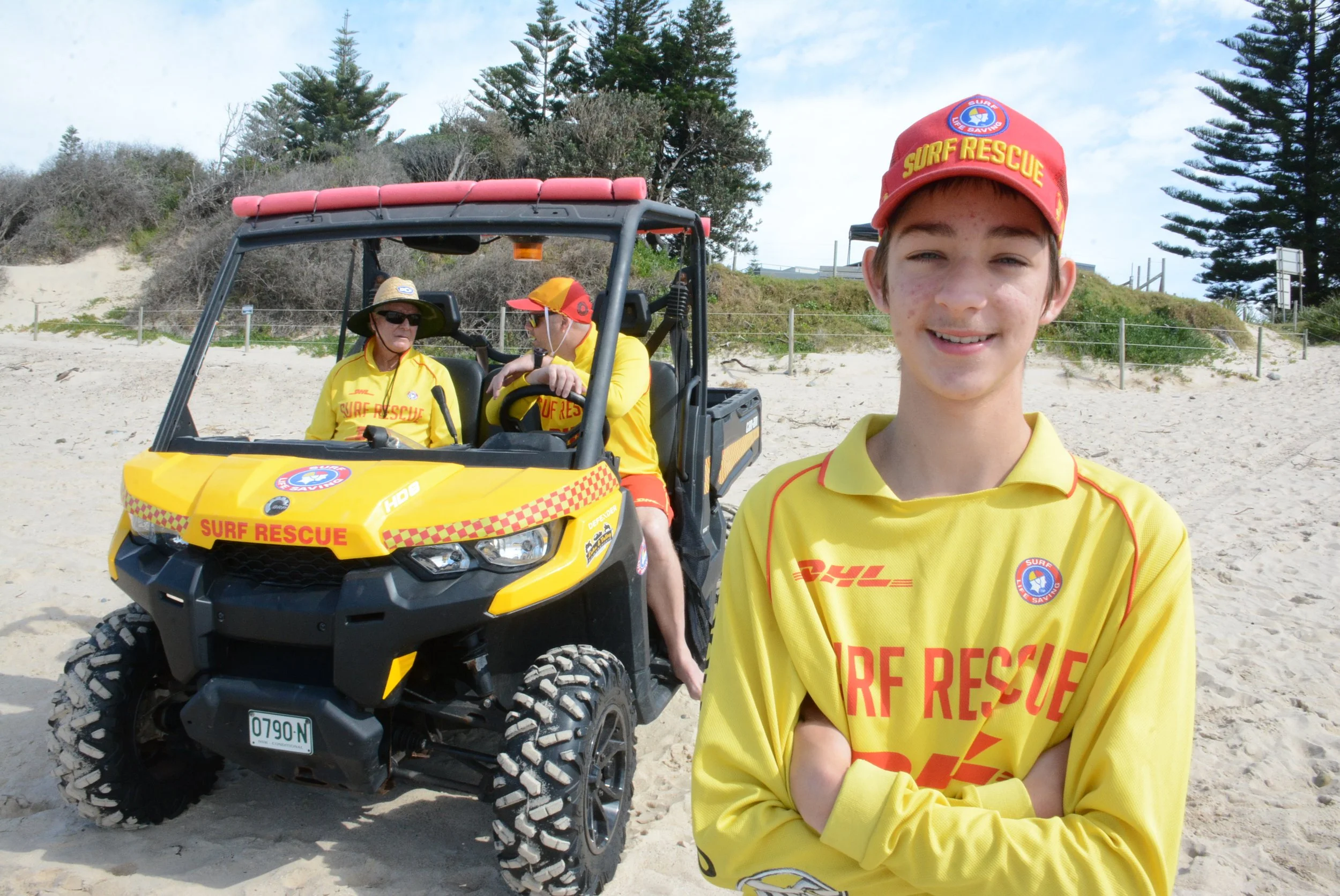 A young boy in a yellow surf rescue shirt and red cap with 'Surf Rescue' written on it, smiling on the beach in front of a yellow off-road rescue vehicle with two adults in yellow shirts and hats, and trees in the background.