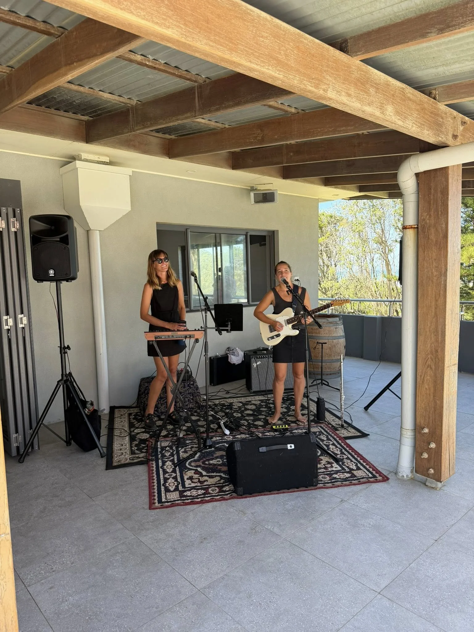 Two women performing music on an outdoor patio with a rug, speakers, microphone, and instruments, including a keyboard and an electric guitar, against a background of trees and a cloudy sky.