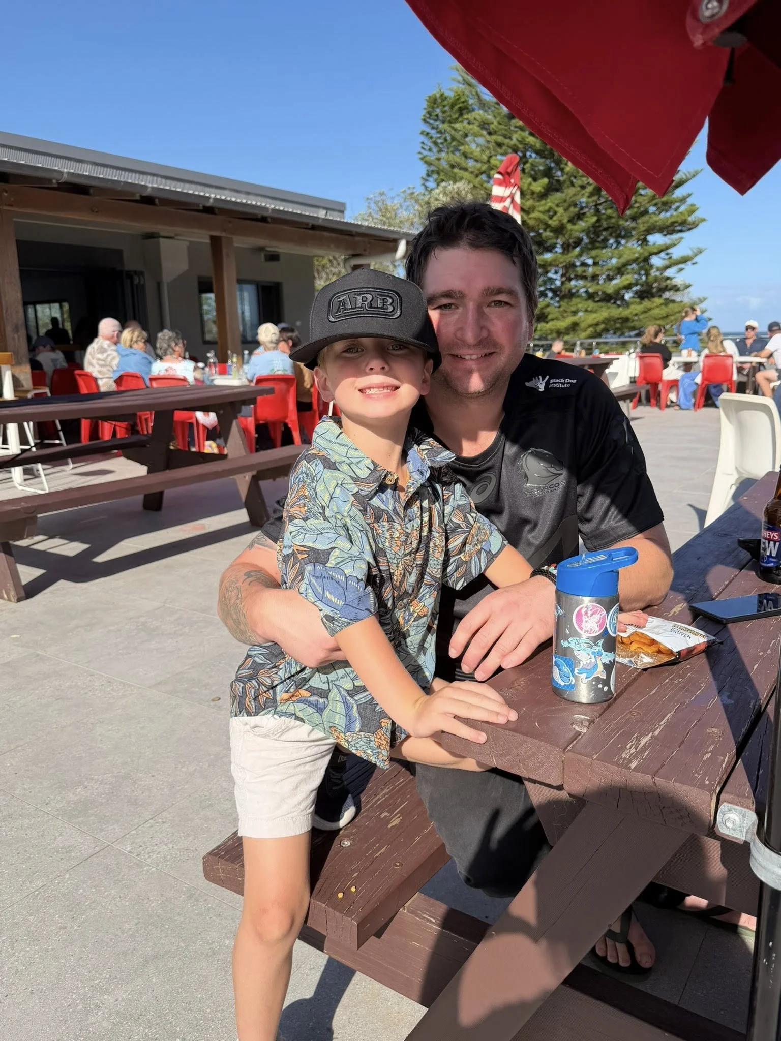 A father and son, smiling at a picnic table outdoors on a sunny day. The boy wears a hat and floral shirt, the man has short dark hair and wears a black shirt. 