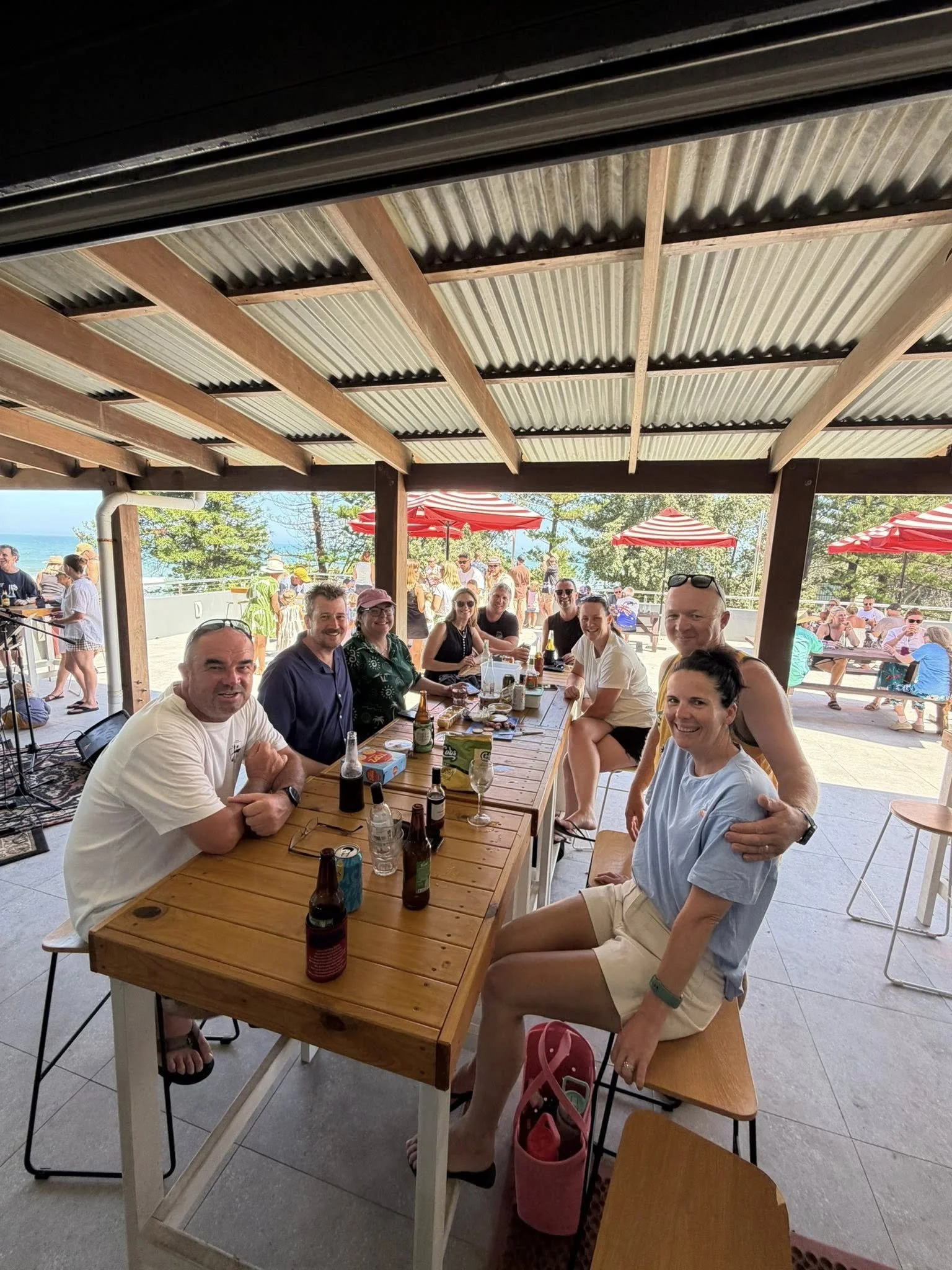 A group of people sitting and enjoying a meal outdoors at a wooden table, with red striped umbrellas and a view of the beach in the background.