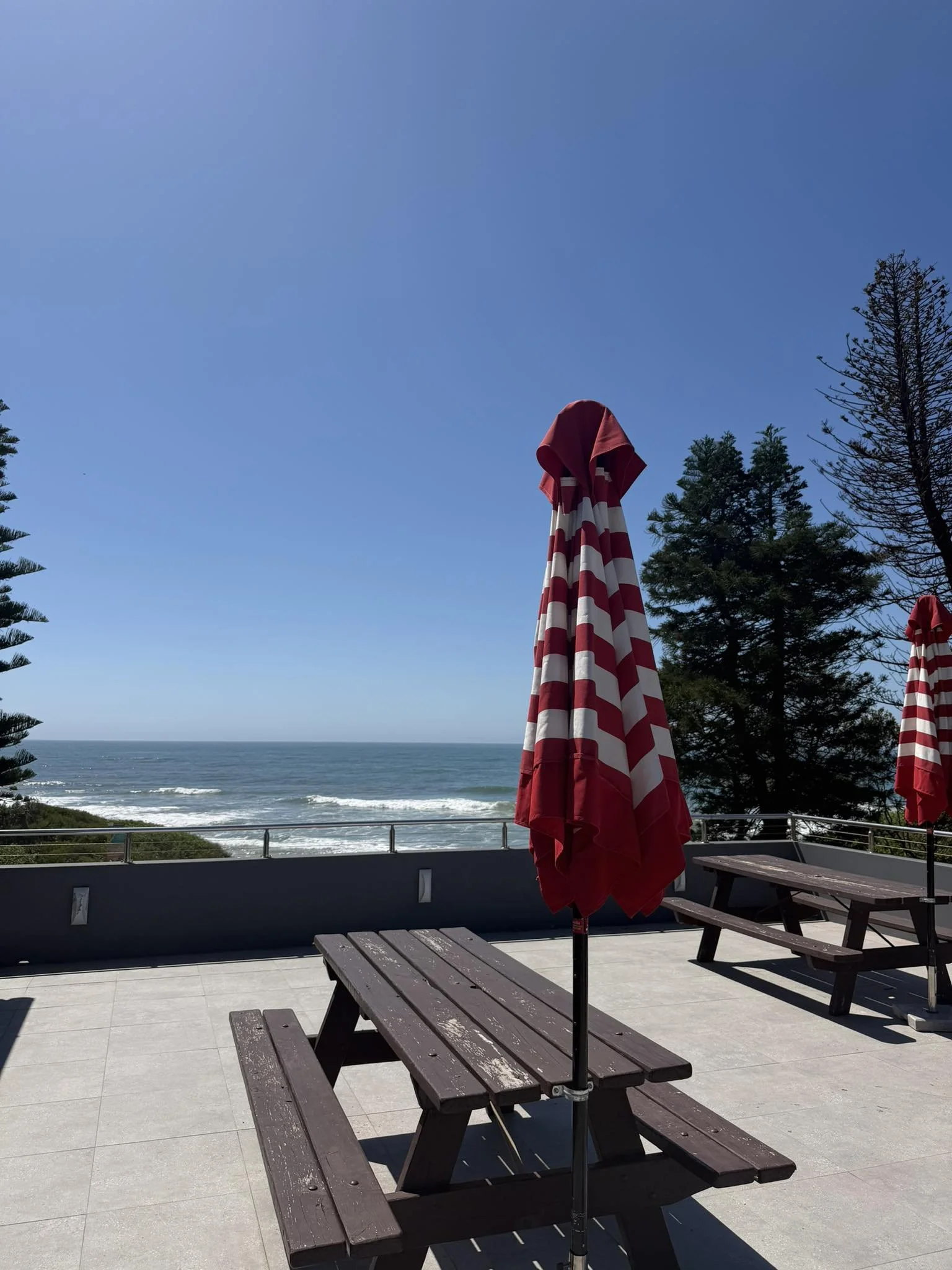 Outdoor patio at the Surf Club with picnic tables and red-and-white striped umbrellas, overlooking the ocean with trees and a clear blue sky.