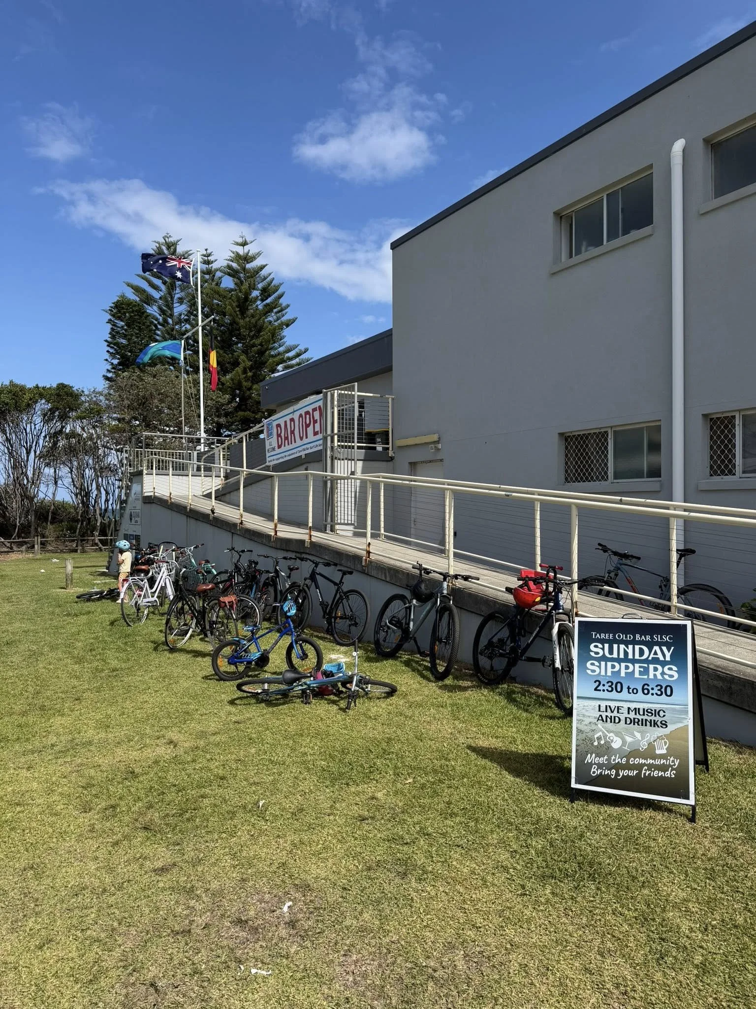 Outside view of the Surf Club with a sign for Sunday Sippers event, featuring live music and drinks. Several bicycles are parked on a grassy area, and flags are flying on poles nearby. Clear blue sky with a few clouds.