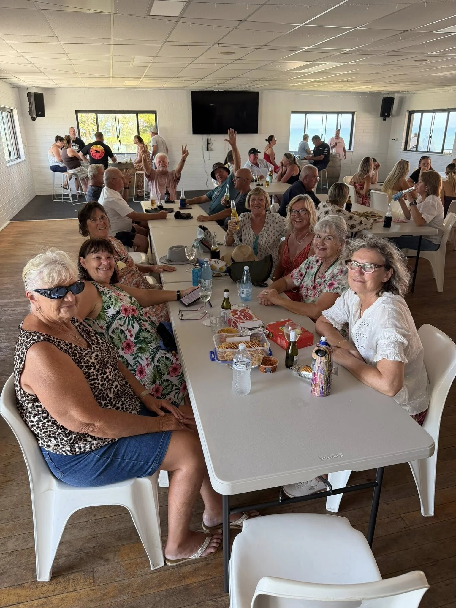 A large gathering of people in a bright room enjoying food and drinks at a social event. Some are sitting at tables, while others are standing and chatting in the background. Many are smiling, waving, and holding beverages. 
