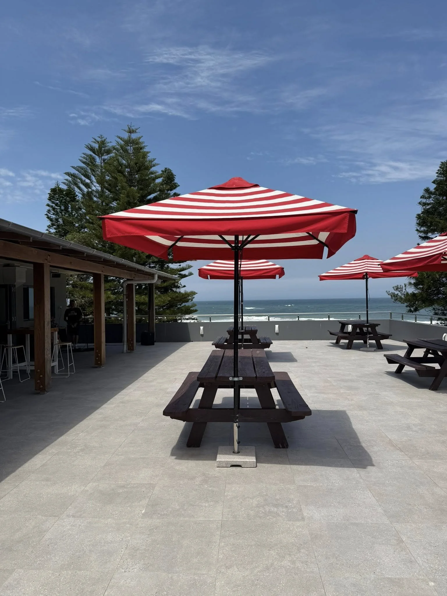 Outdoor patio at the Surf Club with red and white striped umbrellas over wooden picnic tables, ocean view in the background, partly cloudy sky, with trees and a building on the left.