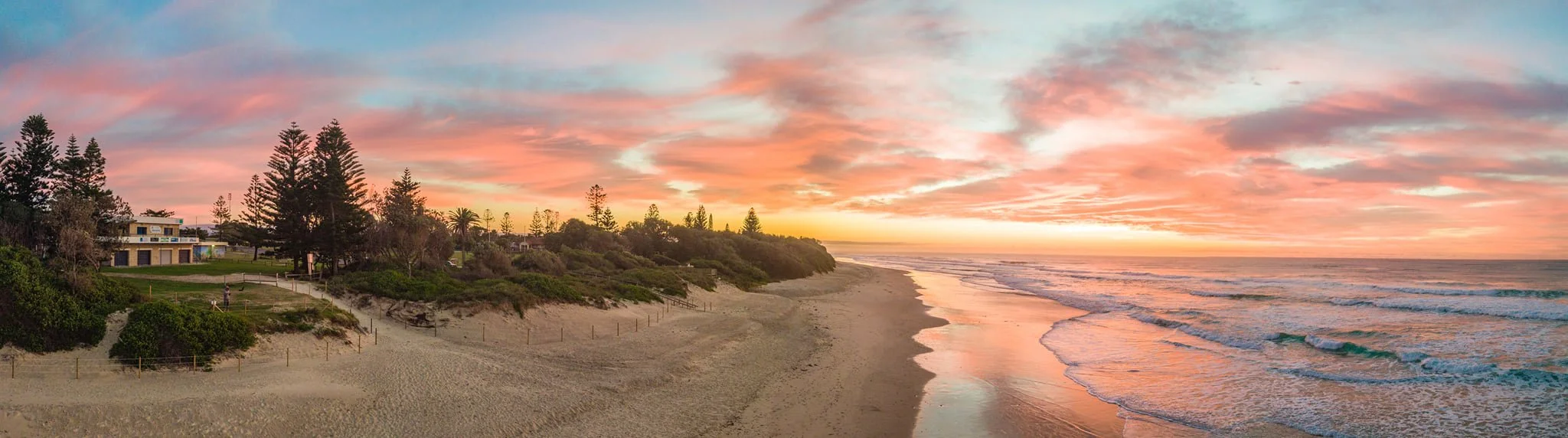 Sunset over a beach with waves, sandy shore, and houses among trees.