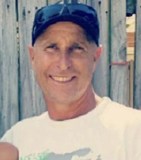 Man smiling outdoors, wearing a blue cap and a white t-shirt, with a wooden fence in the background.