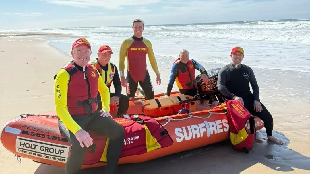 Group of five surf rescue team members in wetsuits and uniforms on a beach with a rescue boat labeled 'SURF RESCUE'