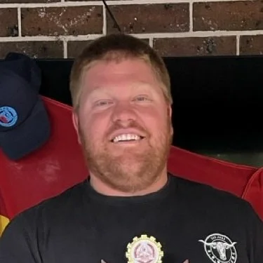 A smiling man with short red hair and a beard sitting outdoors, wearing a black t-shirt with a graphic and a badge, in front of a brick wall.