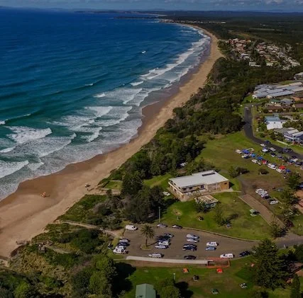 Aerial view of a coastline with a sandy beach, ocean waves, green trees, residential buildings, and parking lots.