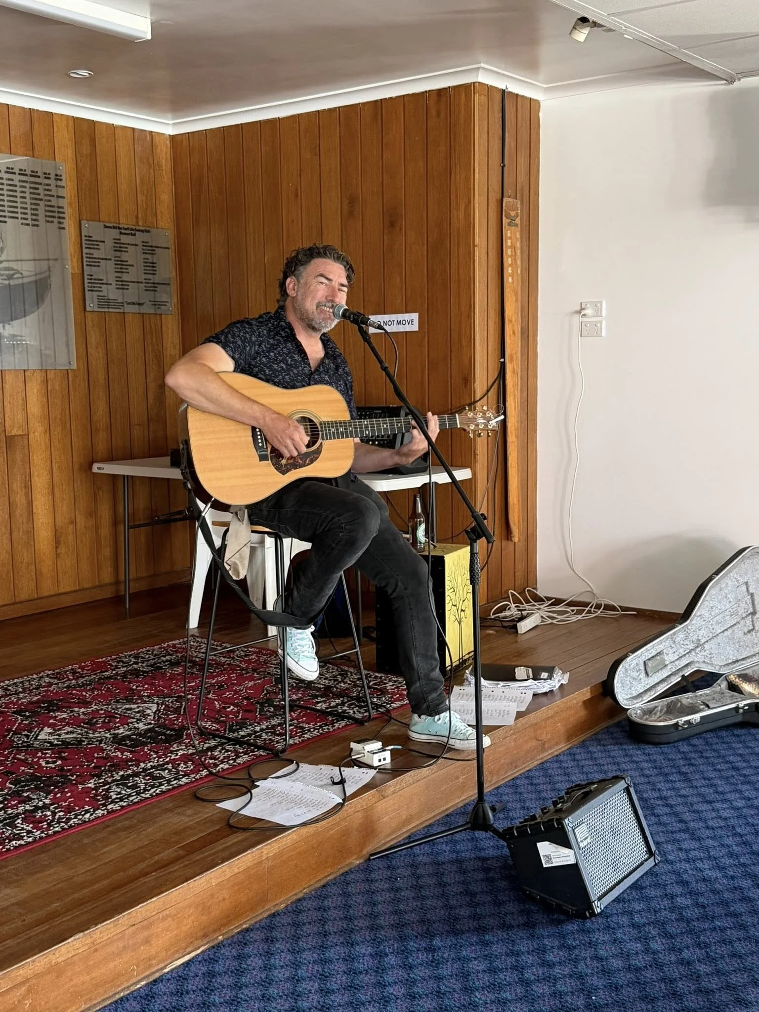 A man with dark hair and a beard, wearing a black patterned shirt and sneakers, is sitting on a stool on a small wooden stage inside the Surf Club. He is playing an acoustic guitar and singing into a microphone.