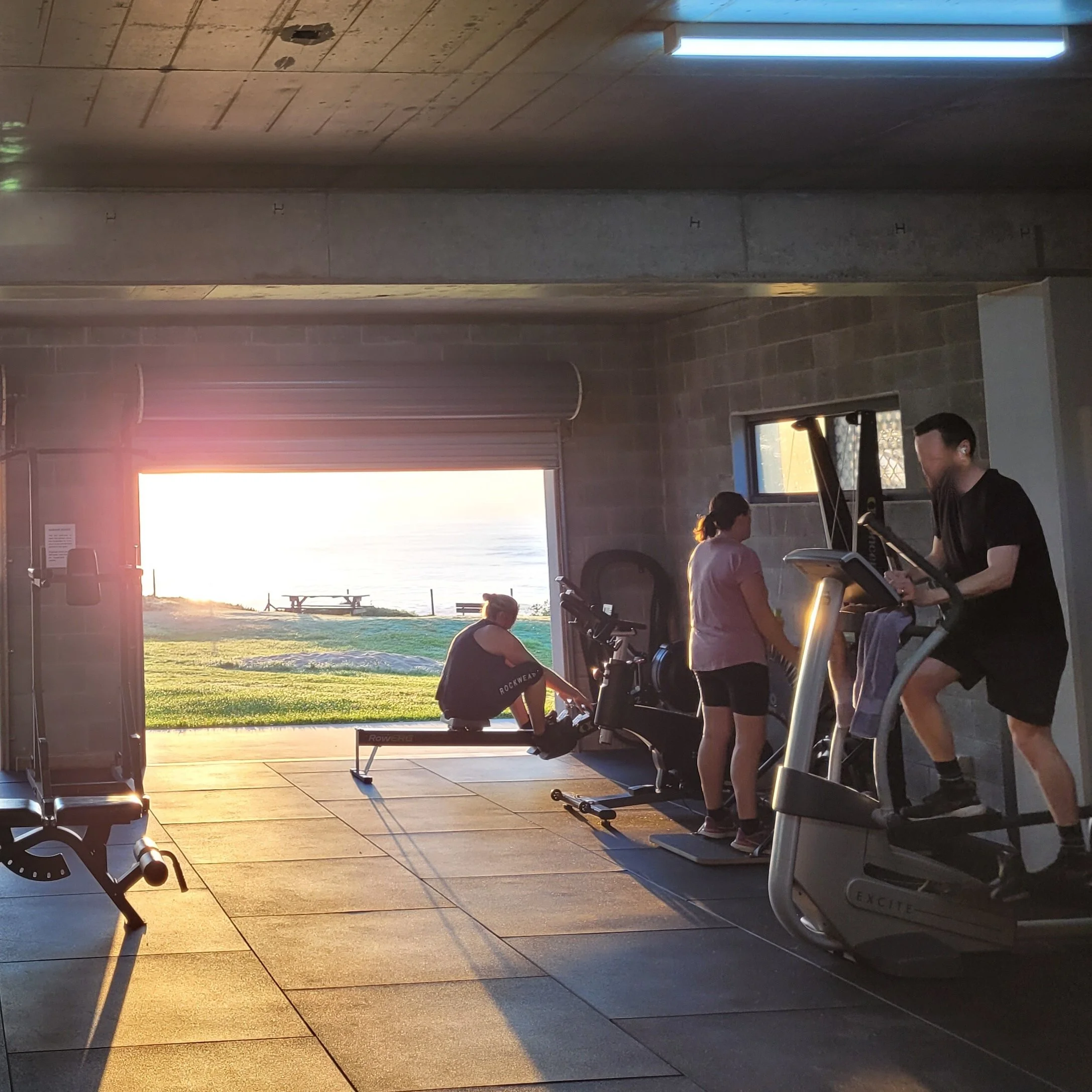 Indoor gym with sunlight streaming through large windows, showing exercise equipment and people working out.