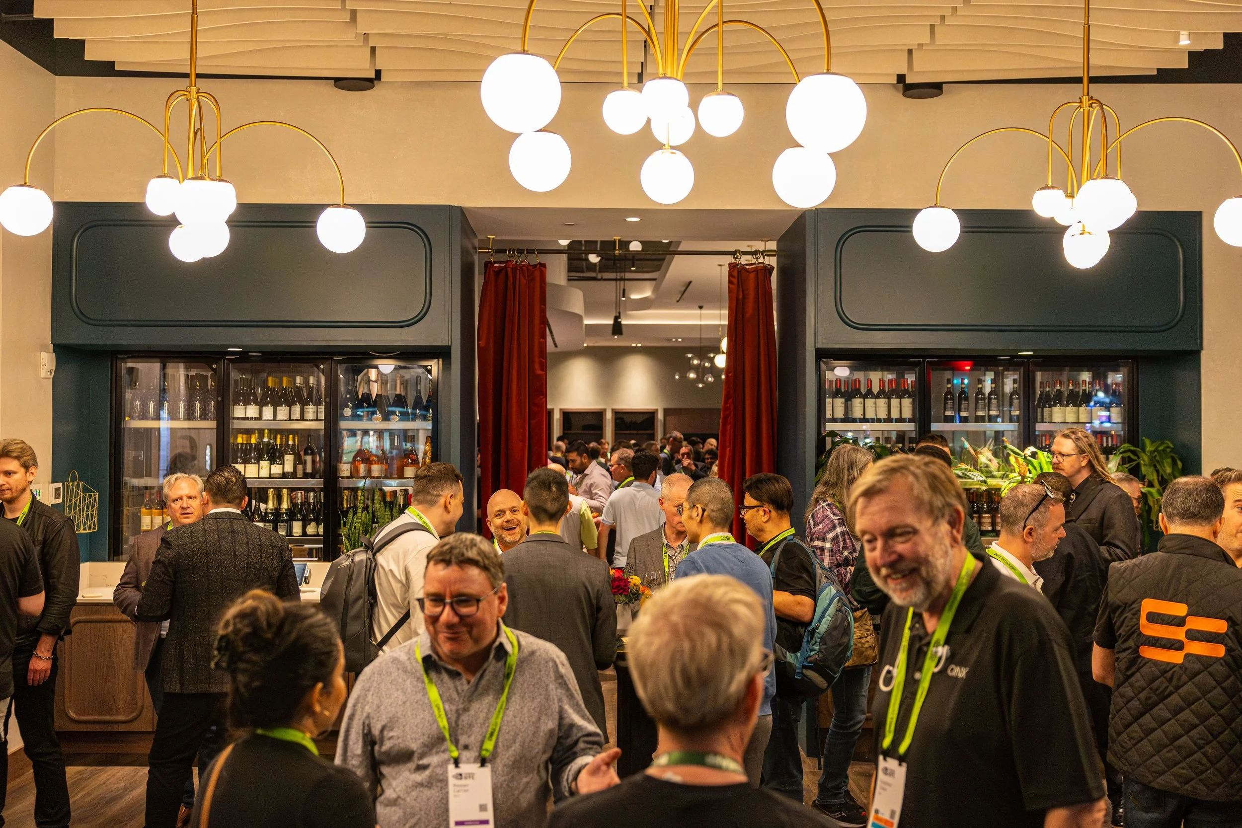 A crowded indoor event with many people mingling, some wearing lanyards and badges. In the background, there is a wine or liquor display with bottles and some red curtains, and a ceiling with modern lighting fixtures including multiple round white gl