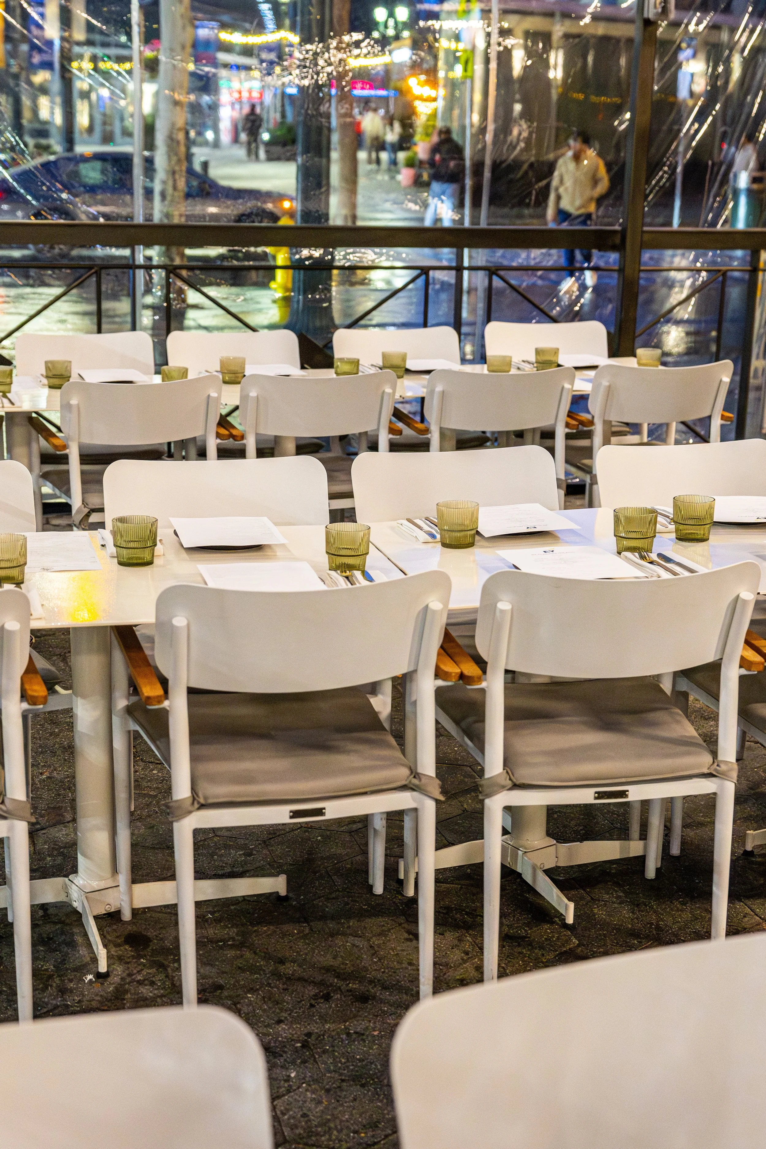 Empty outdoor patio seating area with white chairs and tables, set for dining, and a view through a glass window of a rainy, lively city street with pedestrians and neon signs.