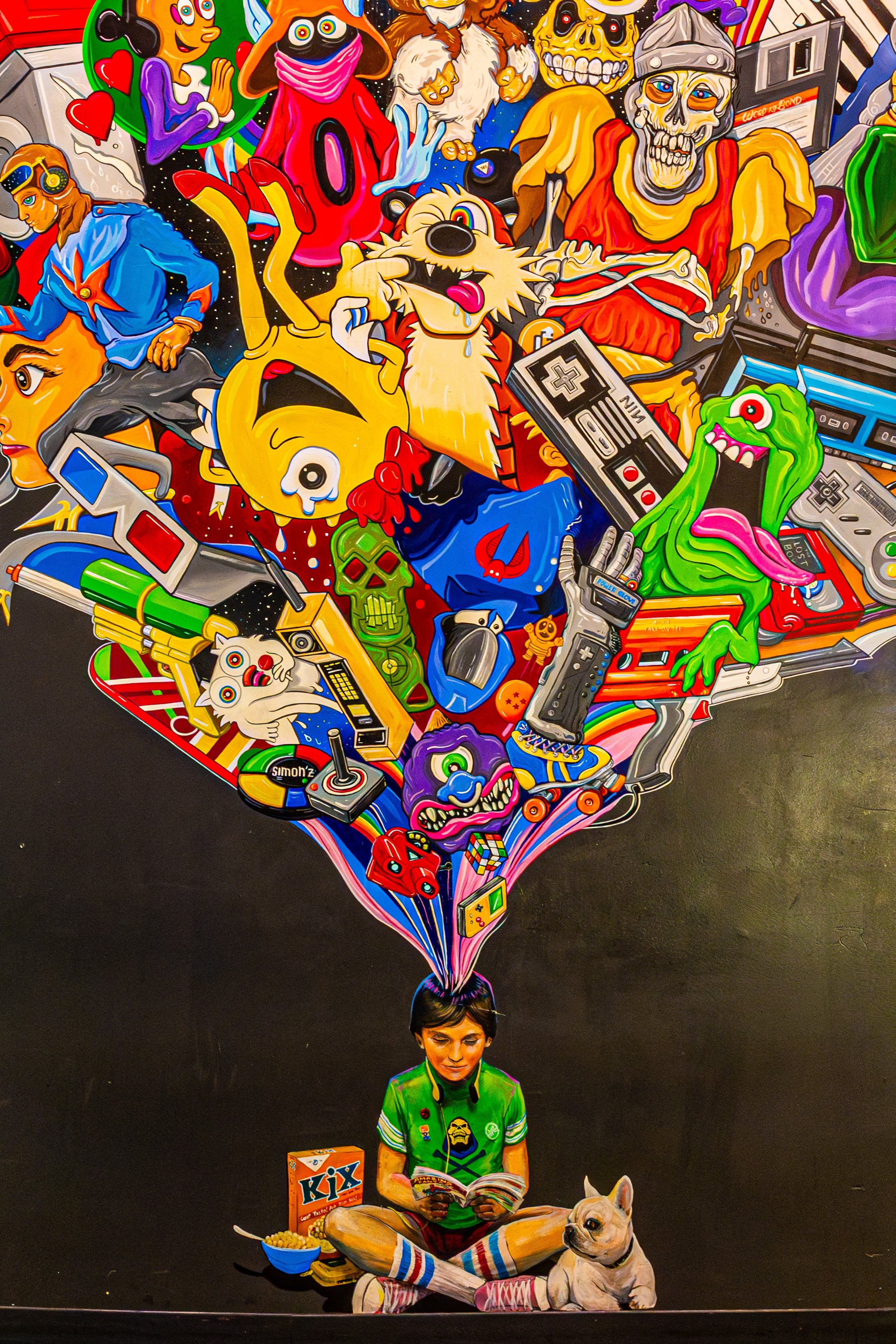 A boy sitting in a cross-legged position, reading a comic book with a Bulldog dog beside him. He is wearing colorful striped socks, shorts, and a green shirt with patches. In front of him is a bowl of popcorn, a box of Kix cereal, and a remote contro