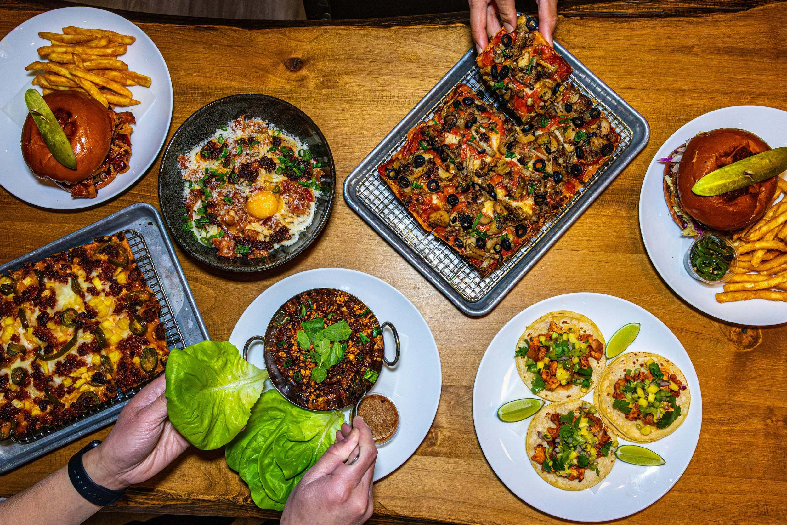 A variety of dishes on a wooden table, including pizza, sliders with pickles, and a person holding lettuce and dipping sauce. The pizza has toppings like pepperoni, pineapple, jalapenos, and ground meat. There are plates with sliders topped with pick