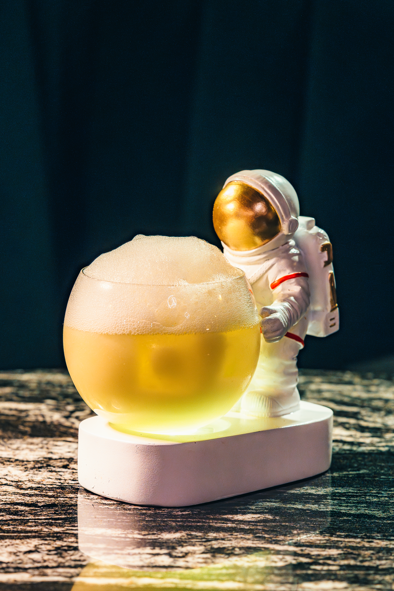 An astronaut figurine in a spacesuit with a reflective gold helmet, standing next to a rounded, glowing yellow object on a white stand, against a dark background with a textured surface beneath.