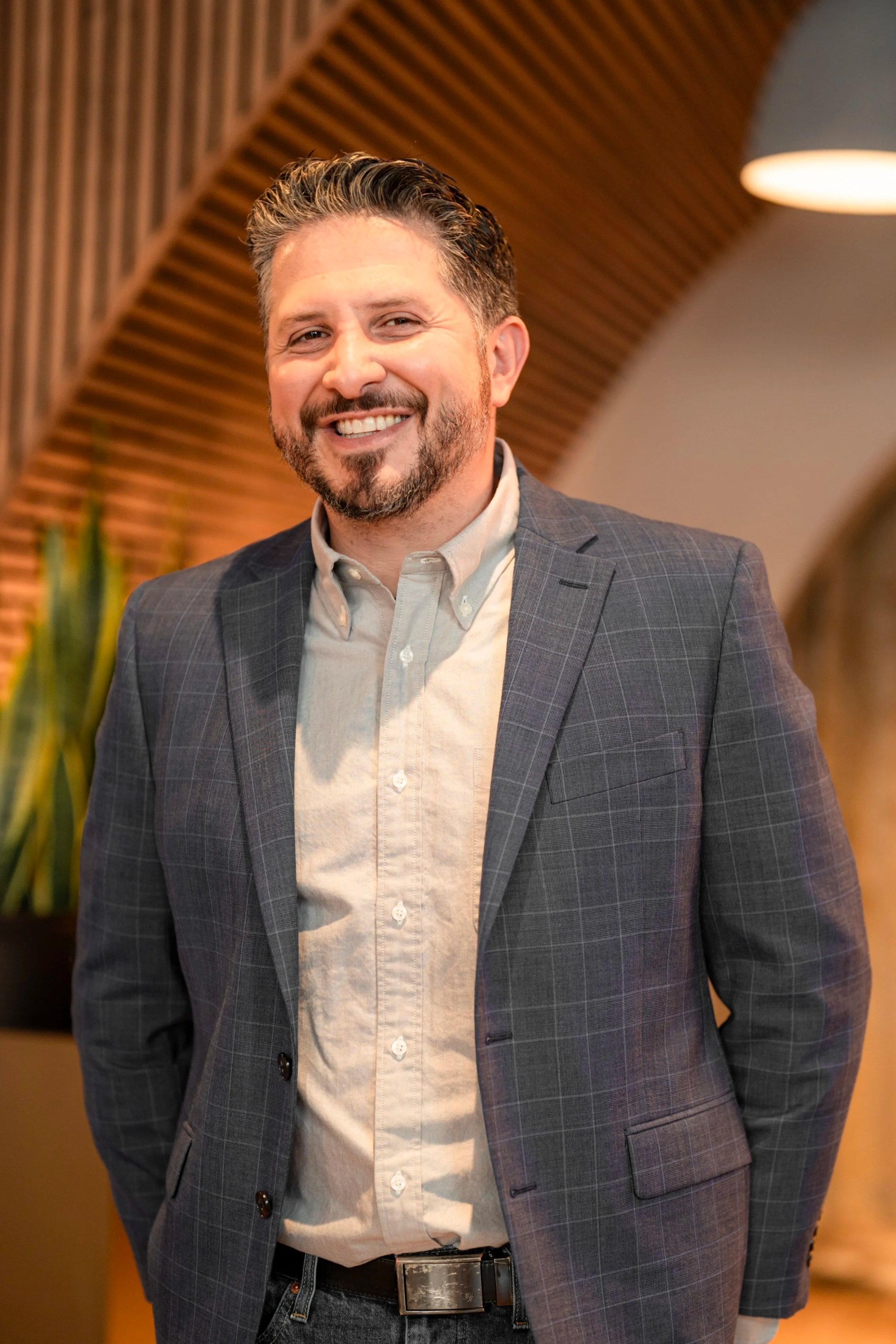 A man with short, styled hair and a beard, smiling and wearing a gray checked blazer over a light button-up shirt, standing indoors with wooden wall decor and a lamp in the background.