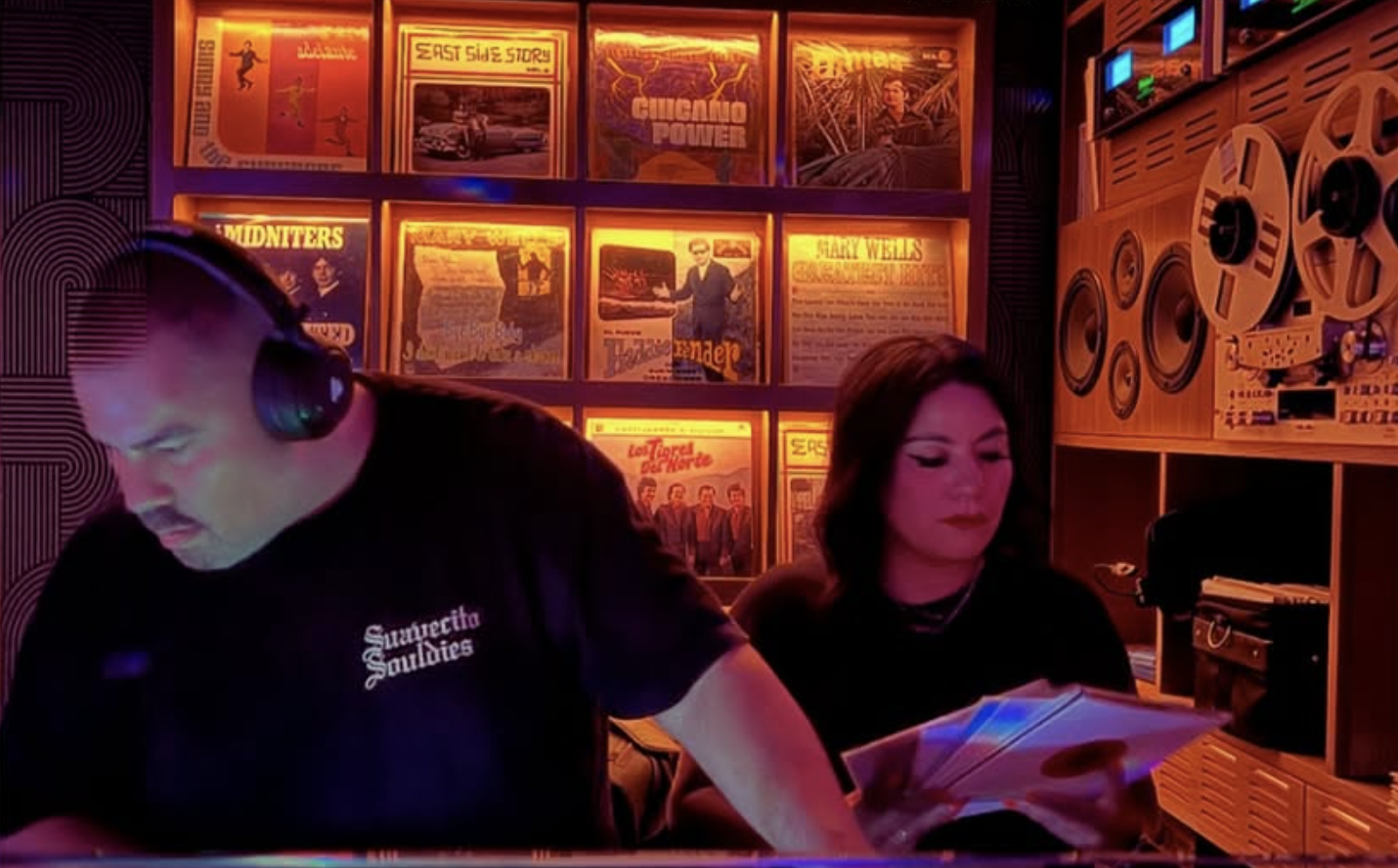 Two people, a man wearing headphones and a woman, are in a dimly lit room with neon lighting. The man is focused on something out of view, and the woman is looking down at a magazine or book. The background features shelves with vinyl records and vintage posters illuminated by orange and purple lights.