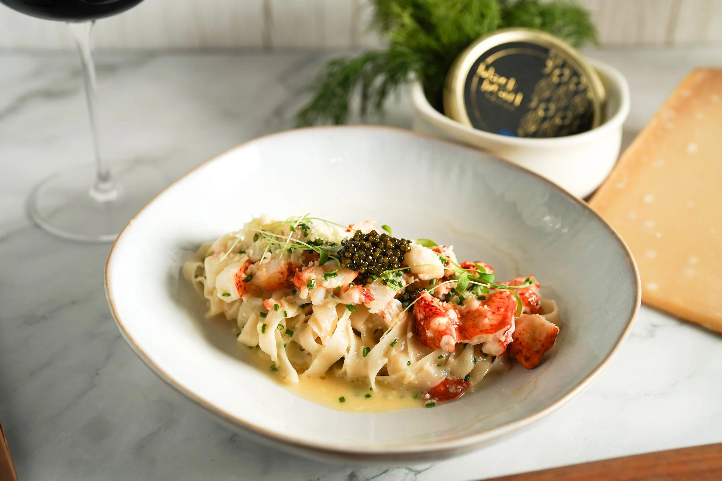 A white bowl of lobster pasta garnished with microgreens and black caviar on top, with a glass of red wine to the side, a wedge of cheese, and a small dish of butter in the background.