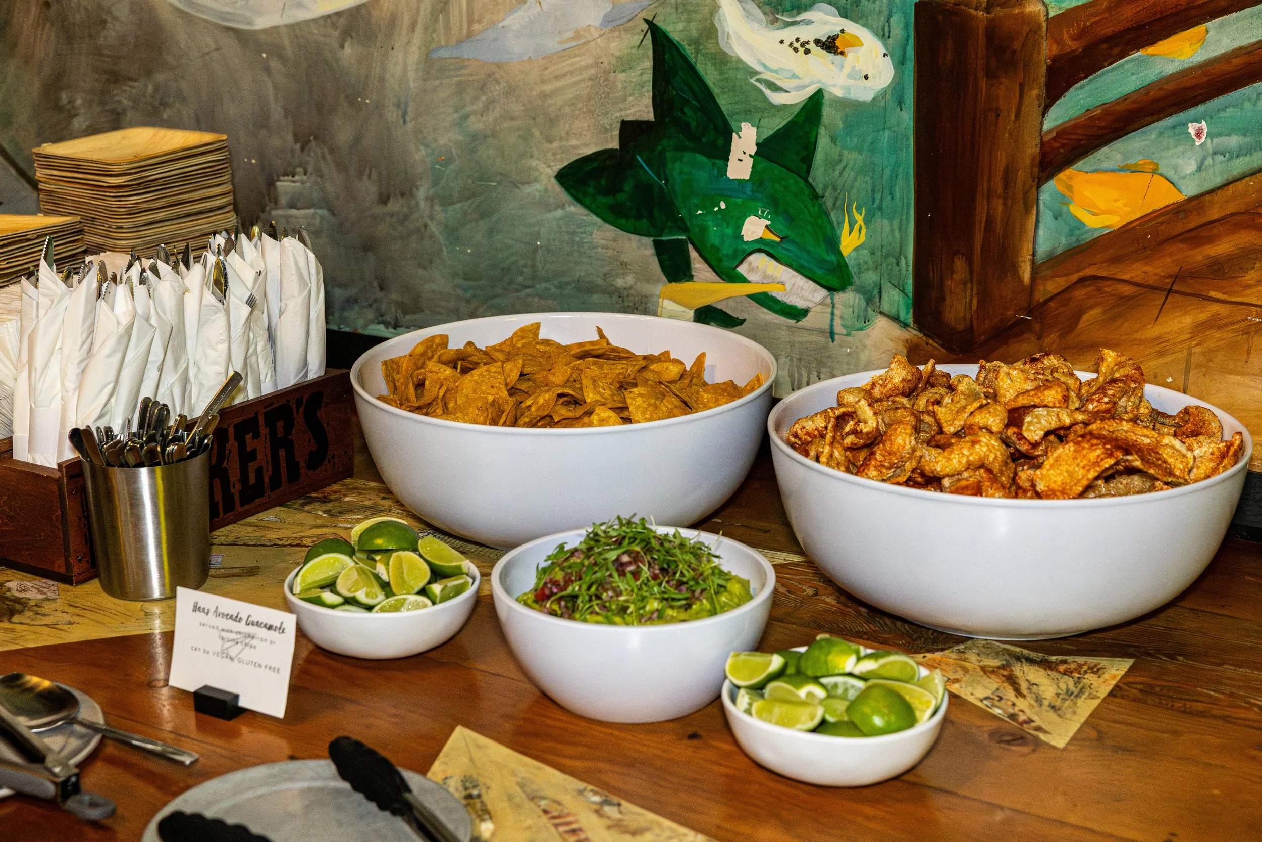 A buffet spread with bowls of chips, fried food, fresh lime wedges, and salad on a wooden table, against a colorful painted wall.