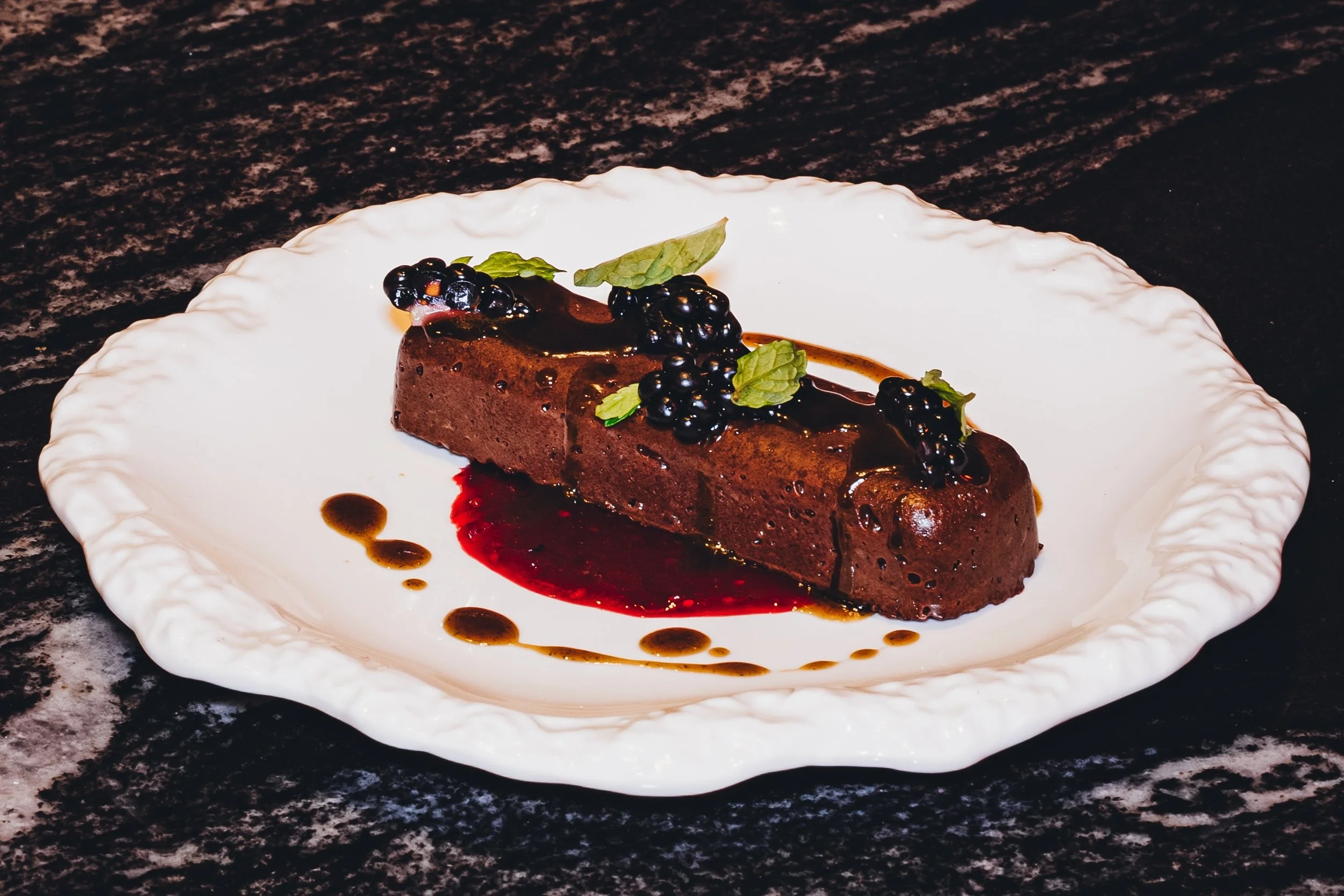 A slice of chocolate mousse cake garnished with blackberries, mint leaves, and drizzled with chocolate and berry sauces on a white, scalloped-edged plate on a dark textured surface.