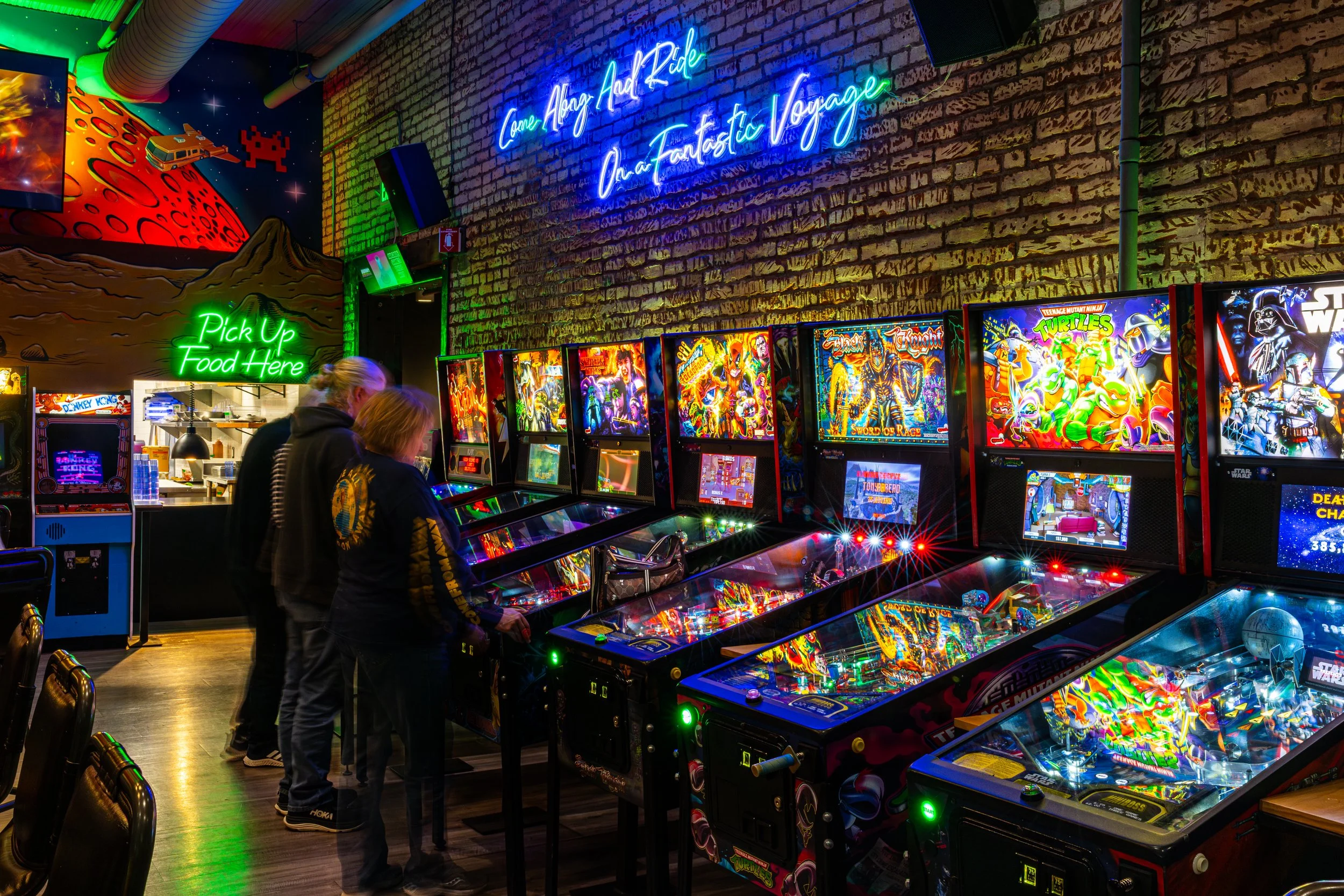 People playing pinball machines in a neon-lit arcade with sci-fi and retro themed decor.