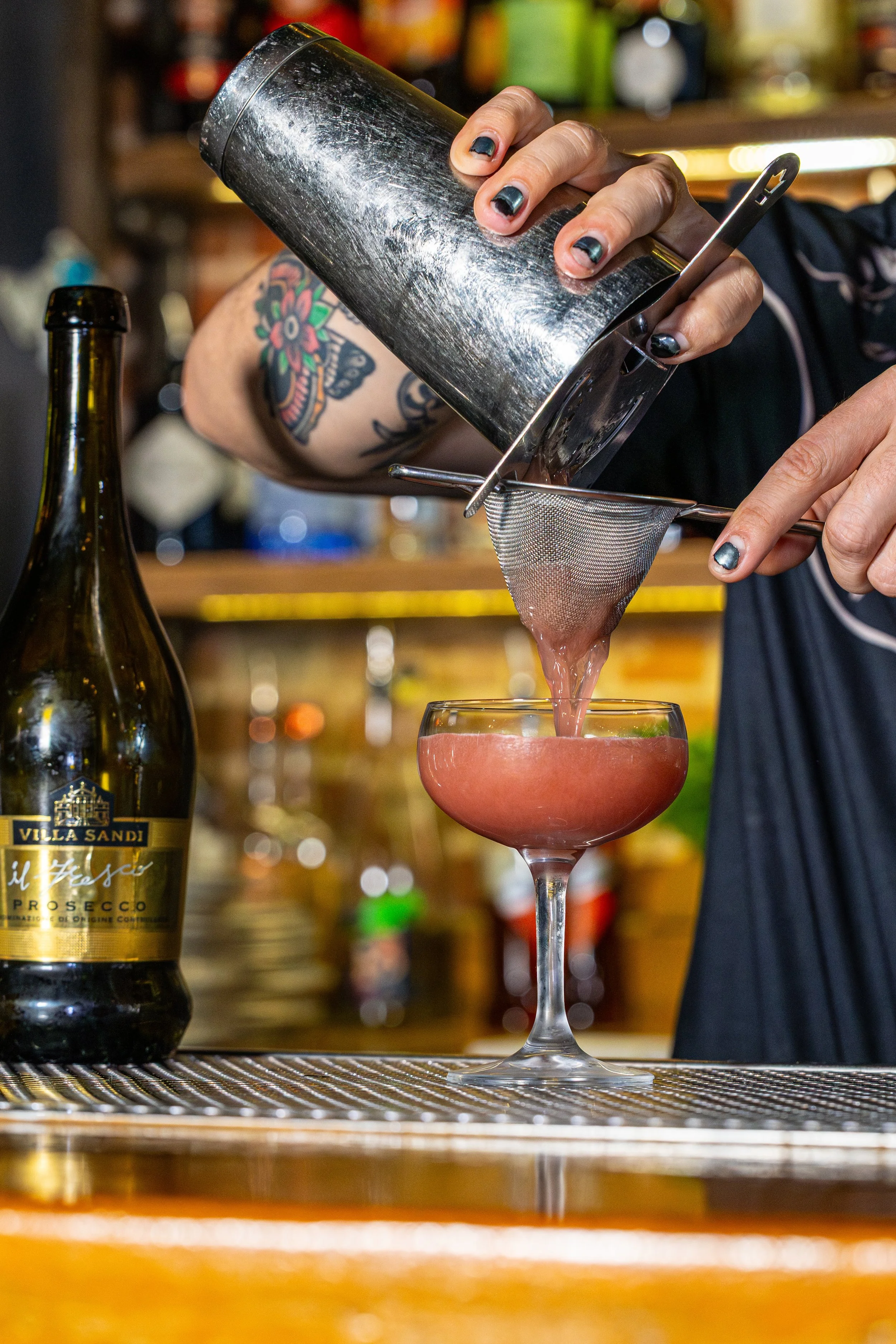A person with colorful tattoos is pouring a pink cocktail through a strainer from a metal shaker into a coupe glass in a bar setting.