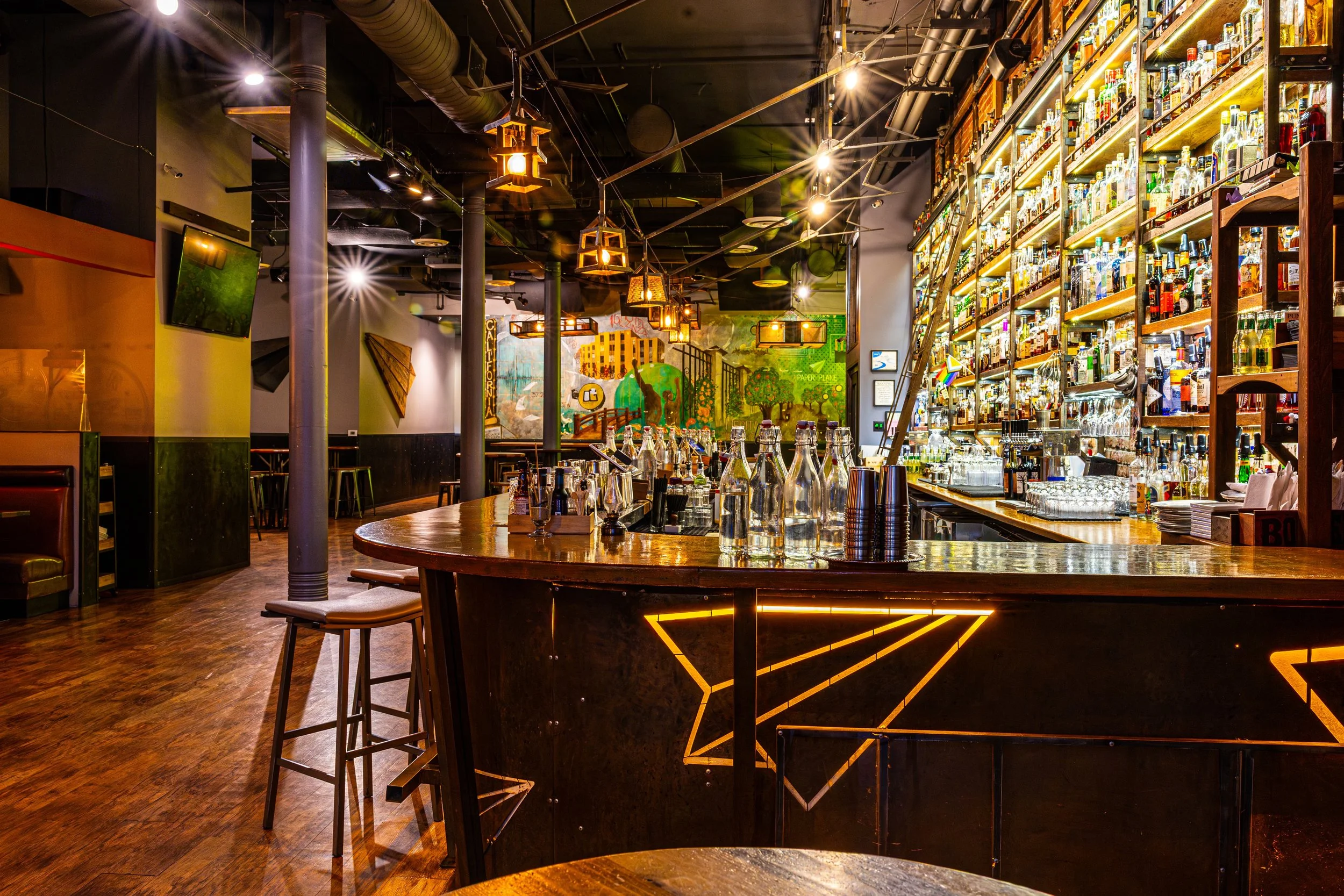 Interior of a stylish bar with wooden tables, bar counter with bottles, modern lighting fixtures, and colorful wall art.