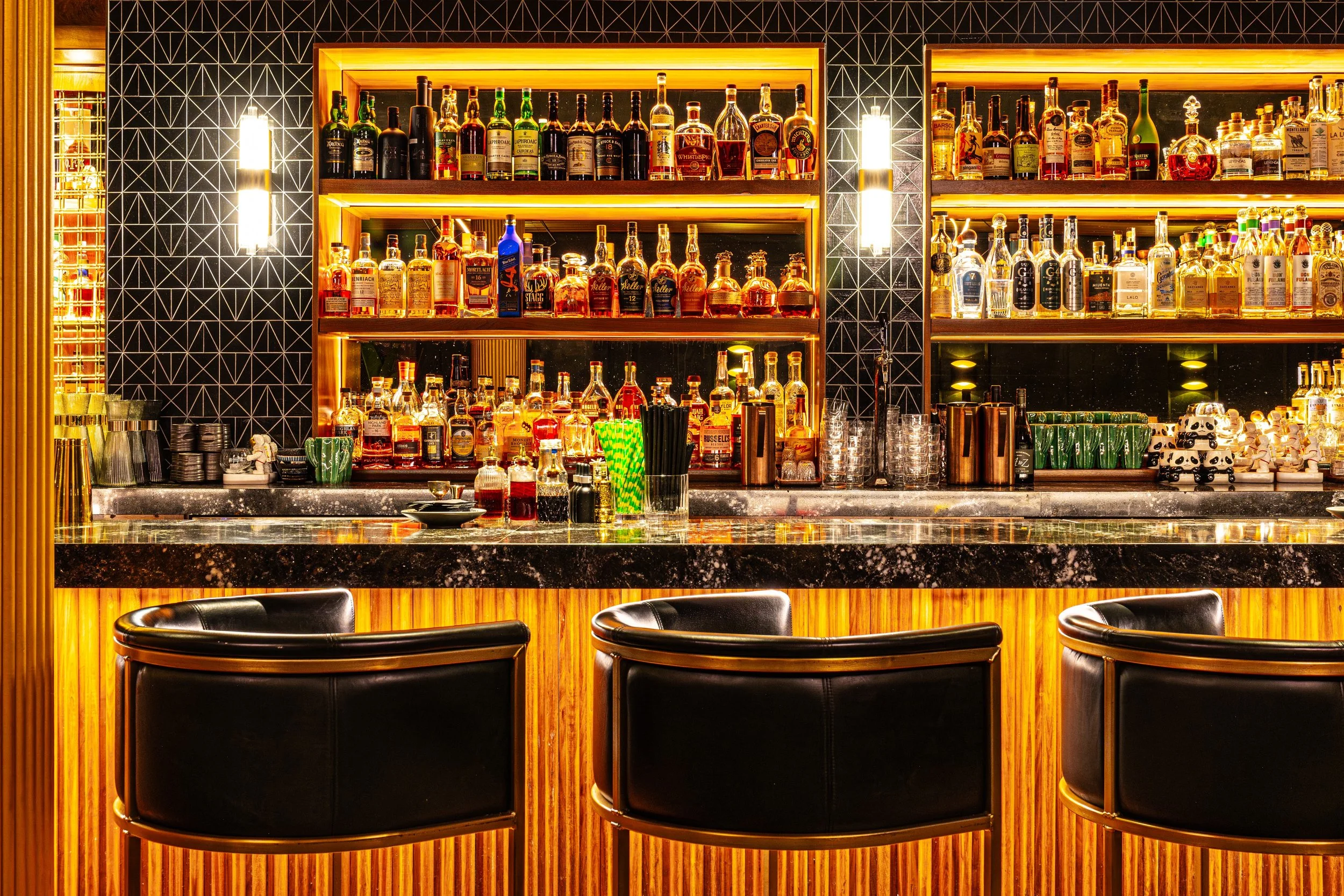 A self-serve bar with wooden shelves filled with various bottles of liquor, illuminated by warm yellow lighting. The counter is black granite, with cocktail stirrers, glasses, and decorative items. Black leather chairs are positioned in front of the bar.