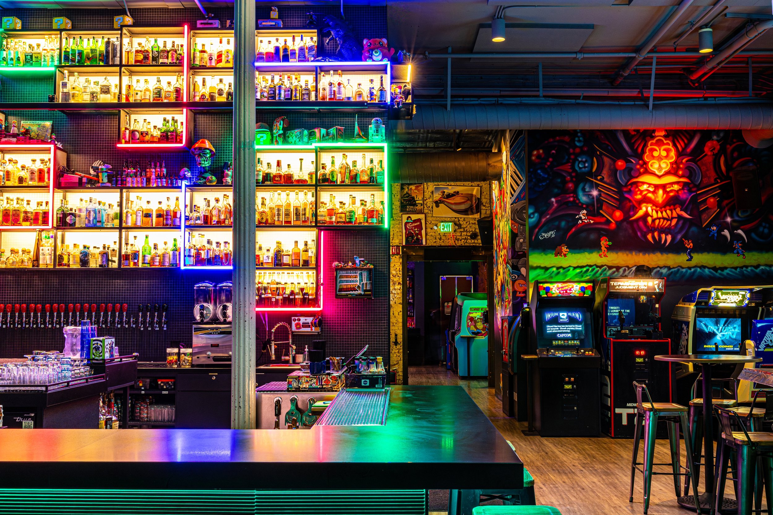 Colorful neon-lit bar area with shelves of bottles, arcade machines, and vibrant wall art.