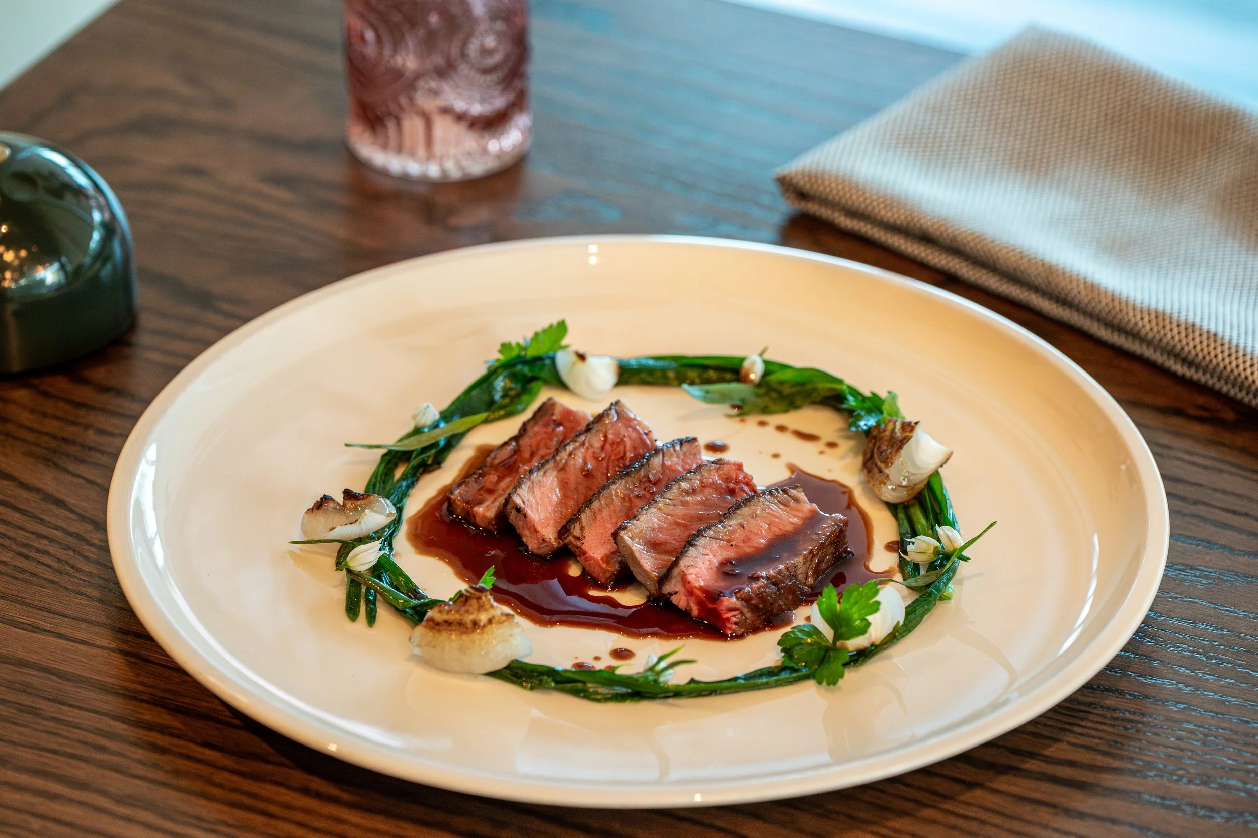 Slices of cooked steak arranged on a white plate, garnished with green herbs and white flowers, with sauce drizzled around.