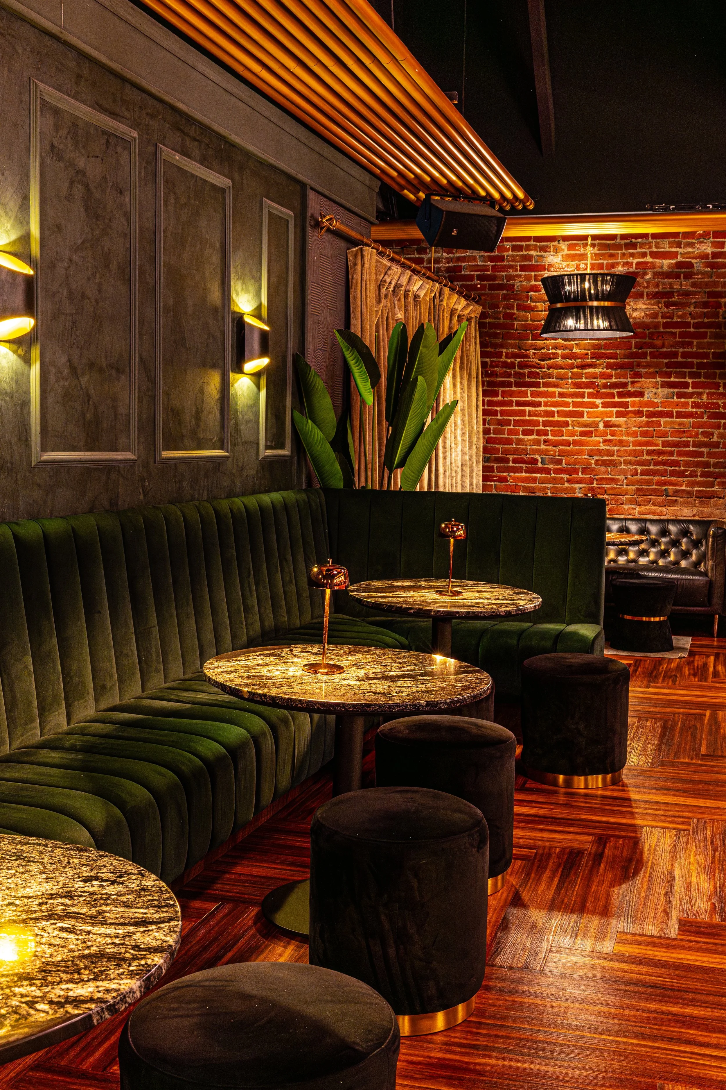 Interior of a stylish bar or lounge with dark green velvet booth seating, round marble-topped tables, black cushioned stools, a brick wall, lush green plants, and warm lighting.