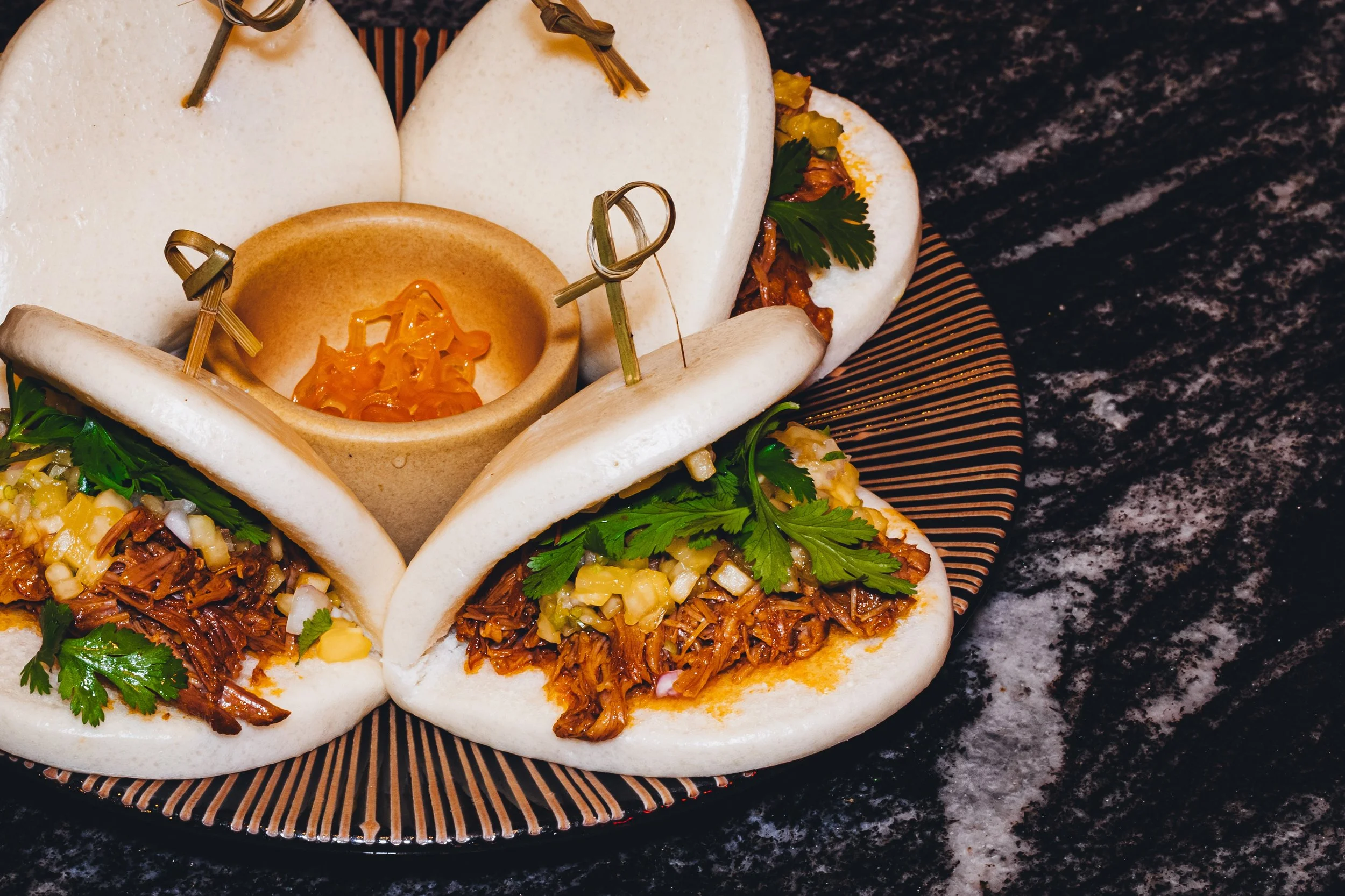 Close-up of several tacos filled with shredded meat, diced onions, chopped cilantro, and topped with jalapenos, served on a decorative black and orange plate on a dark marble countertop.
