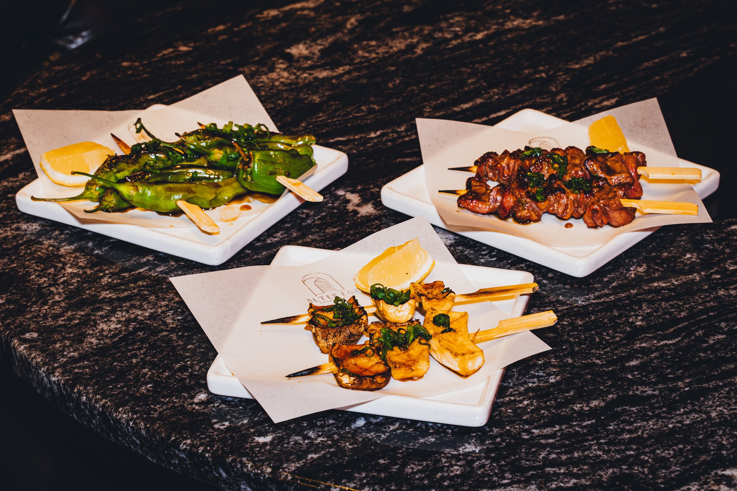 Assorted skewered grilled vegetables and meats on white plates with lemon wedges on a dark textured countertop.