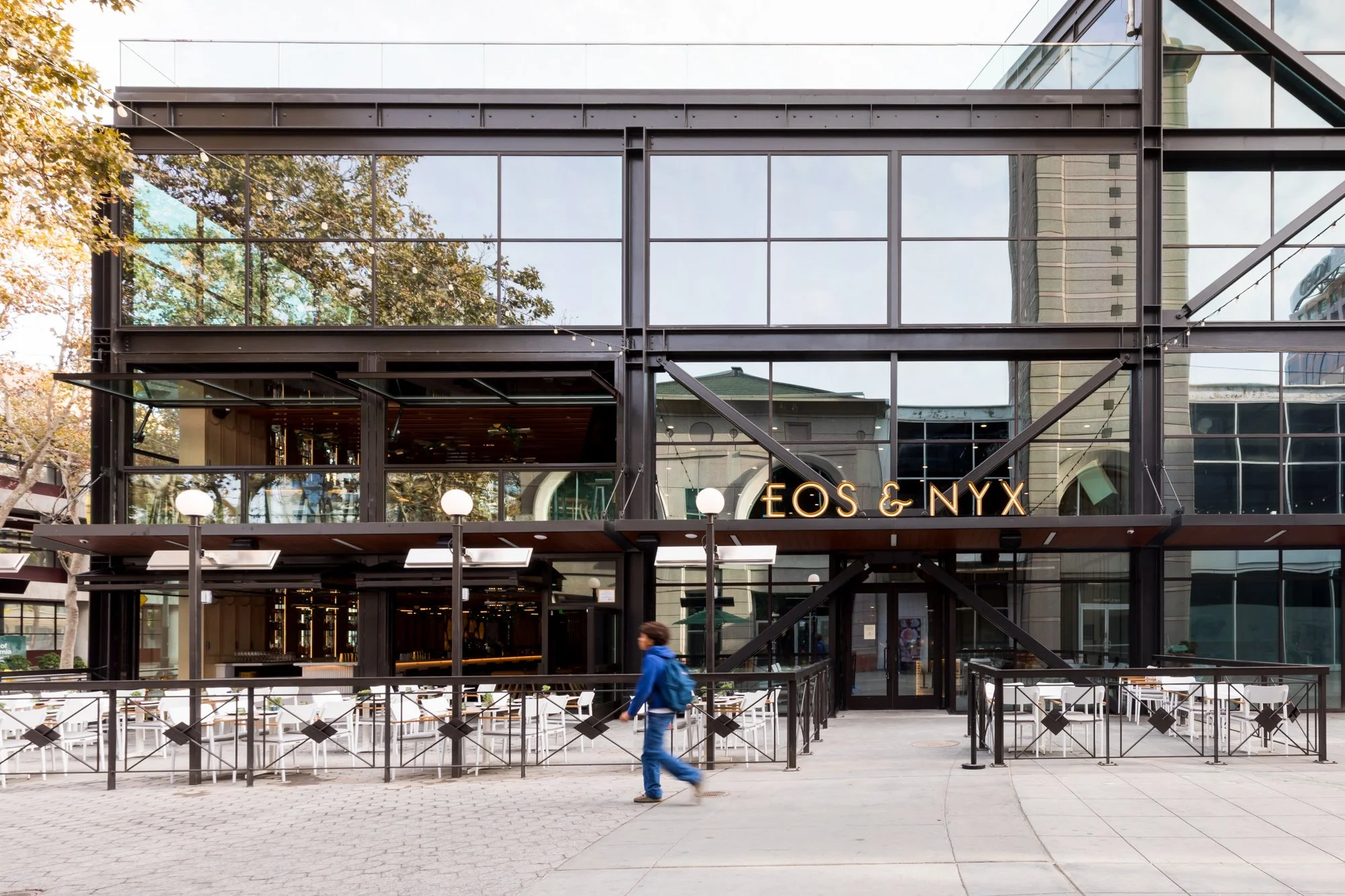 A modern building with large glass windows, a sign that reads 'EOS & NYX', outdoor seating with white chairs, string lights, and a person walking past in a blue jacket.