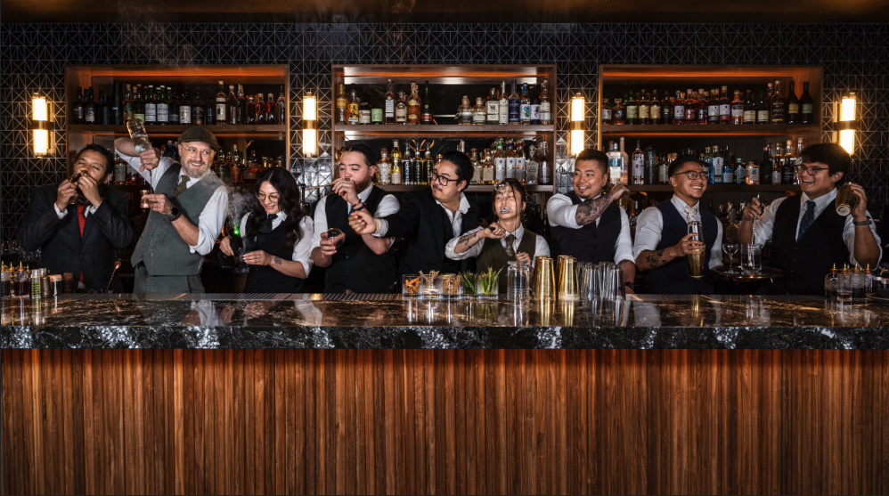 Group of bartenders behind a bar with liquor bottles, smiling and serving drinks.