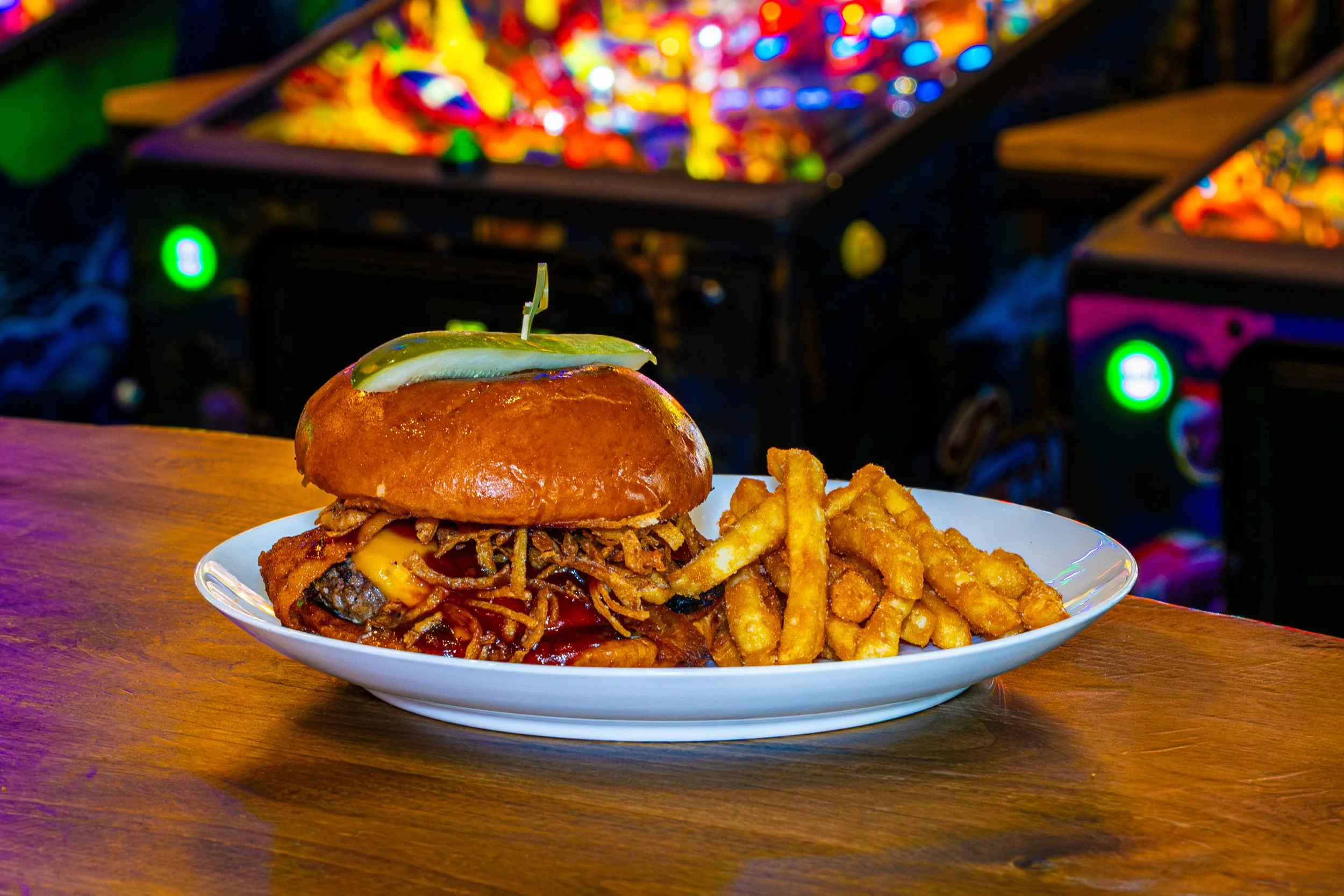 A plate of loaded cheeseburger with crispy fried onion straws and a side of French fries on a wooden table, with colorful, blurred carnival or arcade lights in the background.