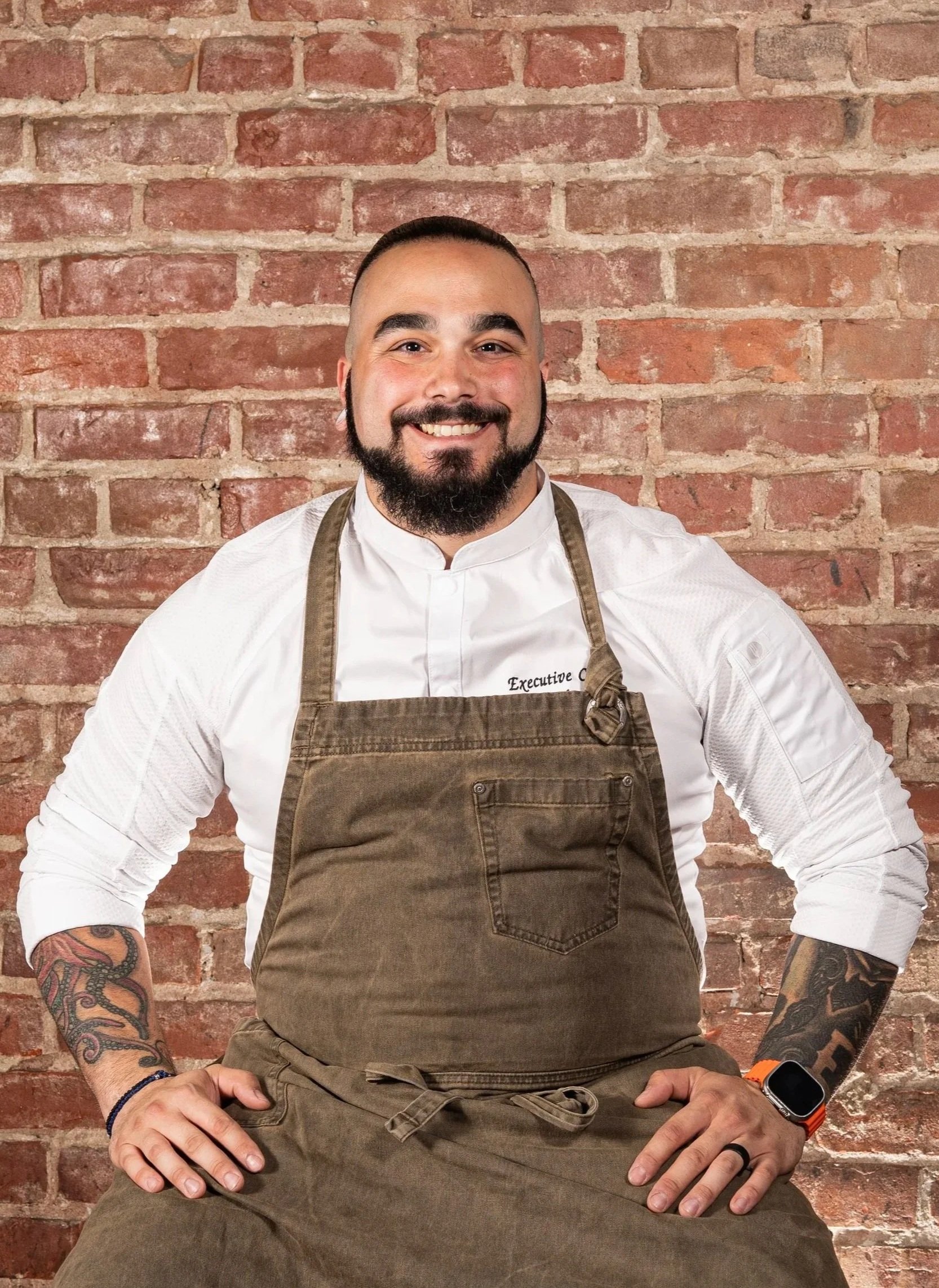 A smiling man with tattoos on his arms wearing a white shirt, brown apron, and an orange wristwatch, sitting in front of a red brick wall.