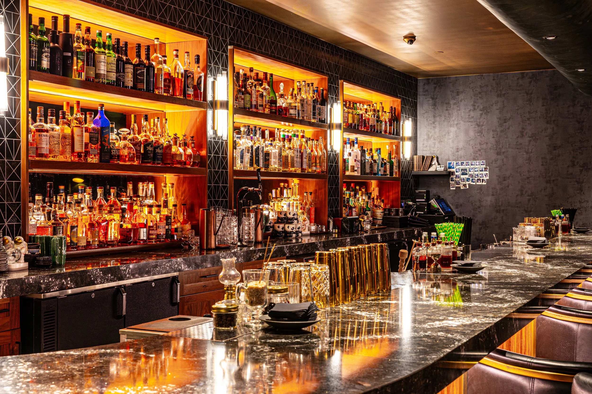 A bar with illuminated wooden shelves stocked with various bottles of liquor, a black marble countertop with barware, and a textured gray wall with a small photo display.