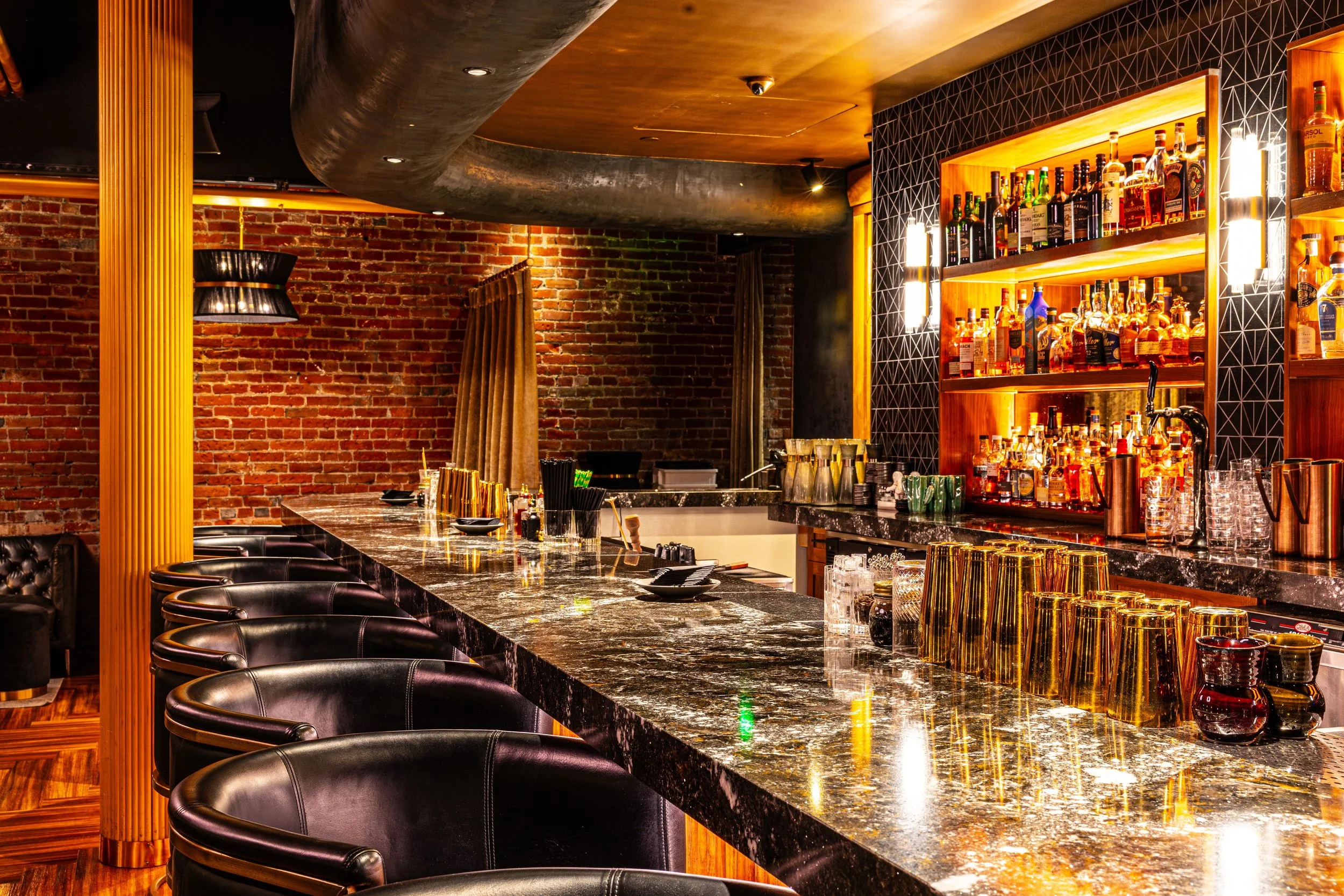A stylish bar with a marble countertop, black leather stools, a backlit wooden shelf with various bottles of alcohol, exposed brick walls, and modern lighting fixtures.
