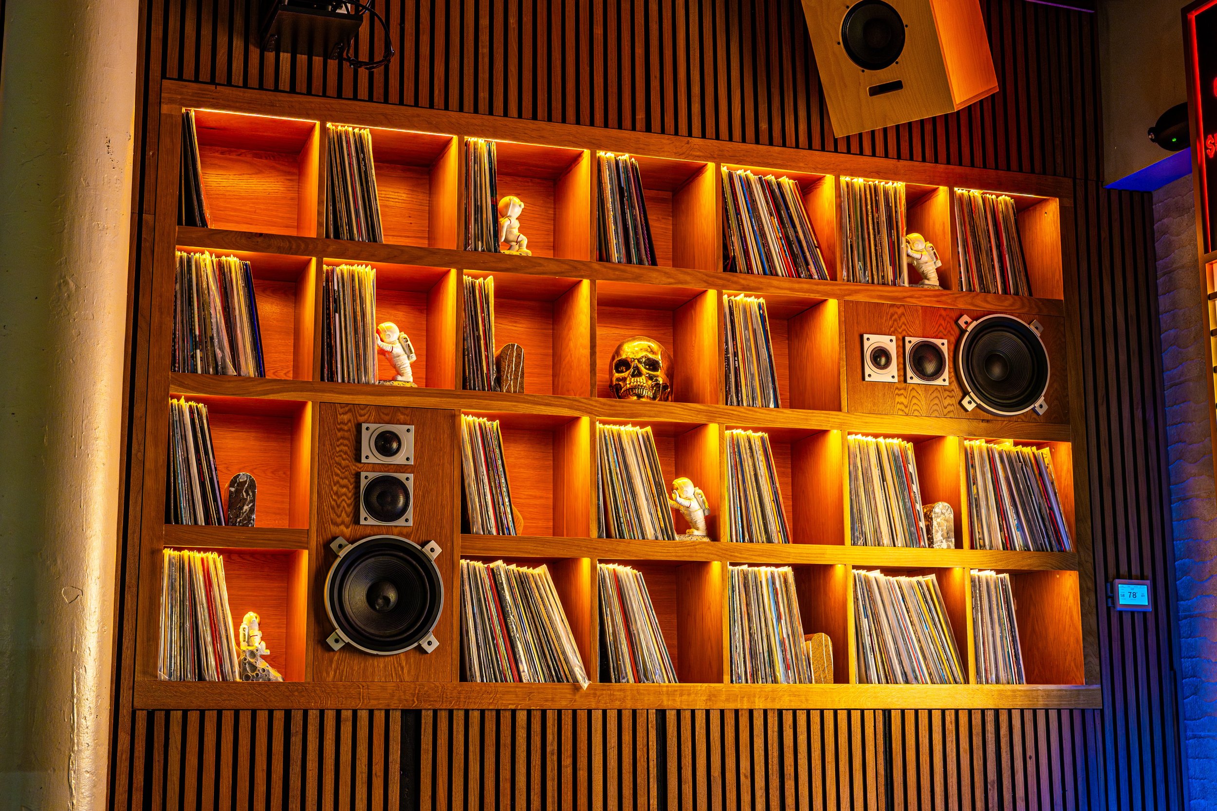 Wooden bookshelf with backlit shelves holding vinyl records, small astronaut figurines, a gold skull, and speakers, in a dimly lit room.