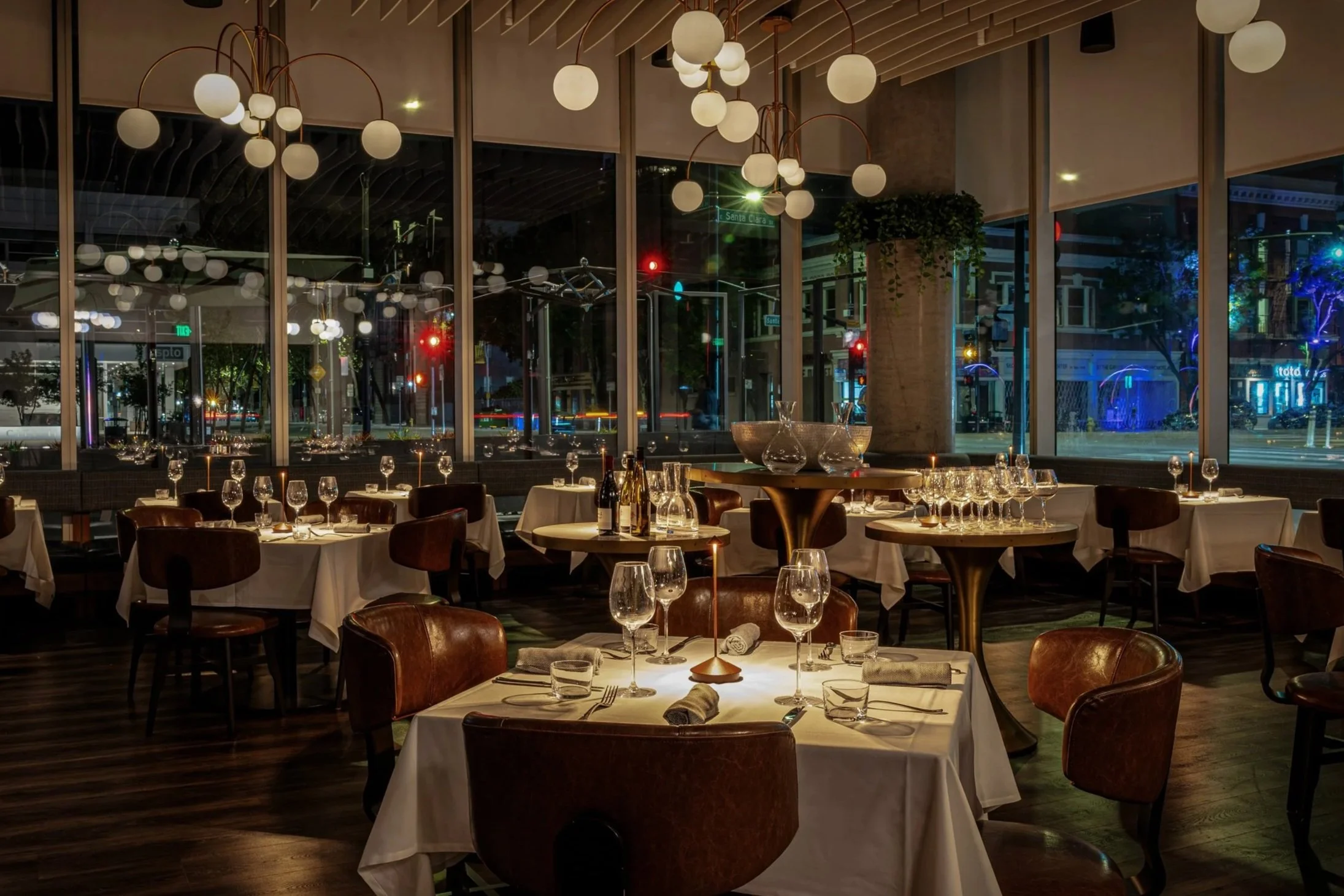 A cozy, elegant restaurant interior with large windows showing city lights outside, modern chandeliers hanging from the ceiling, and tables set with white tablecloths, wine glasses, and napkins.