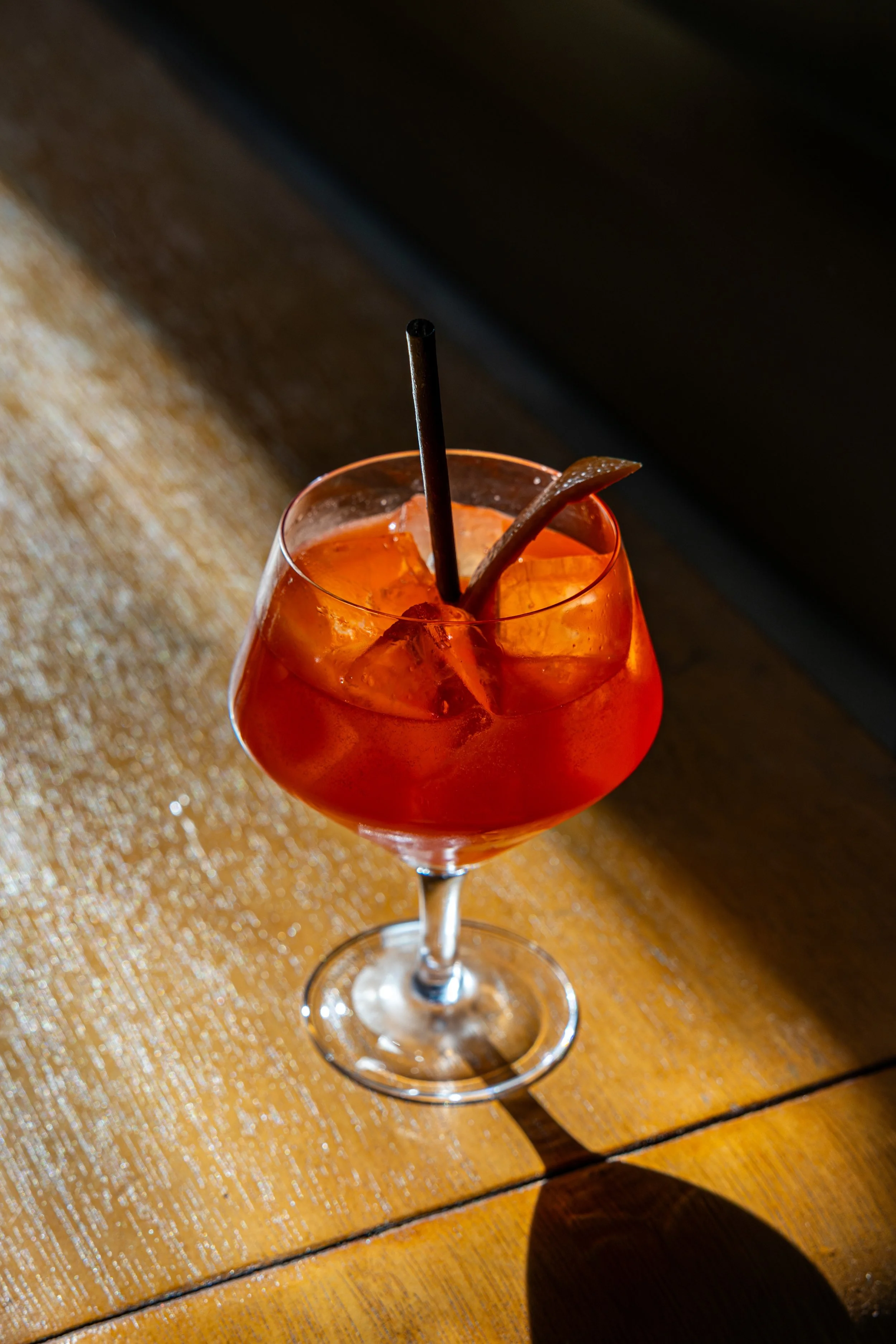 A close-up of a cocktail glass with orange-colored drink and ice, garnished with a slice of fruit and a black straw, on a wooden table.
