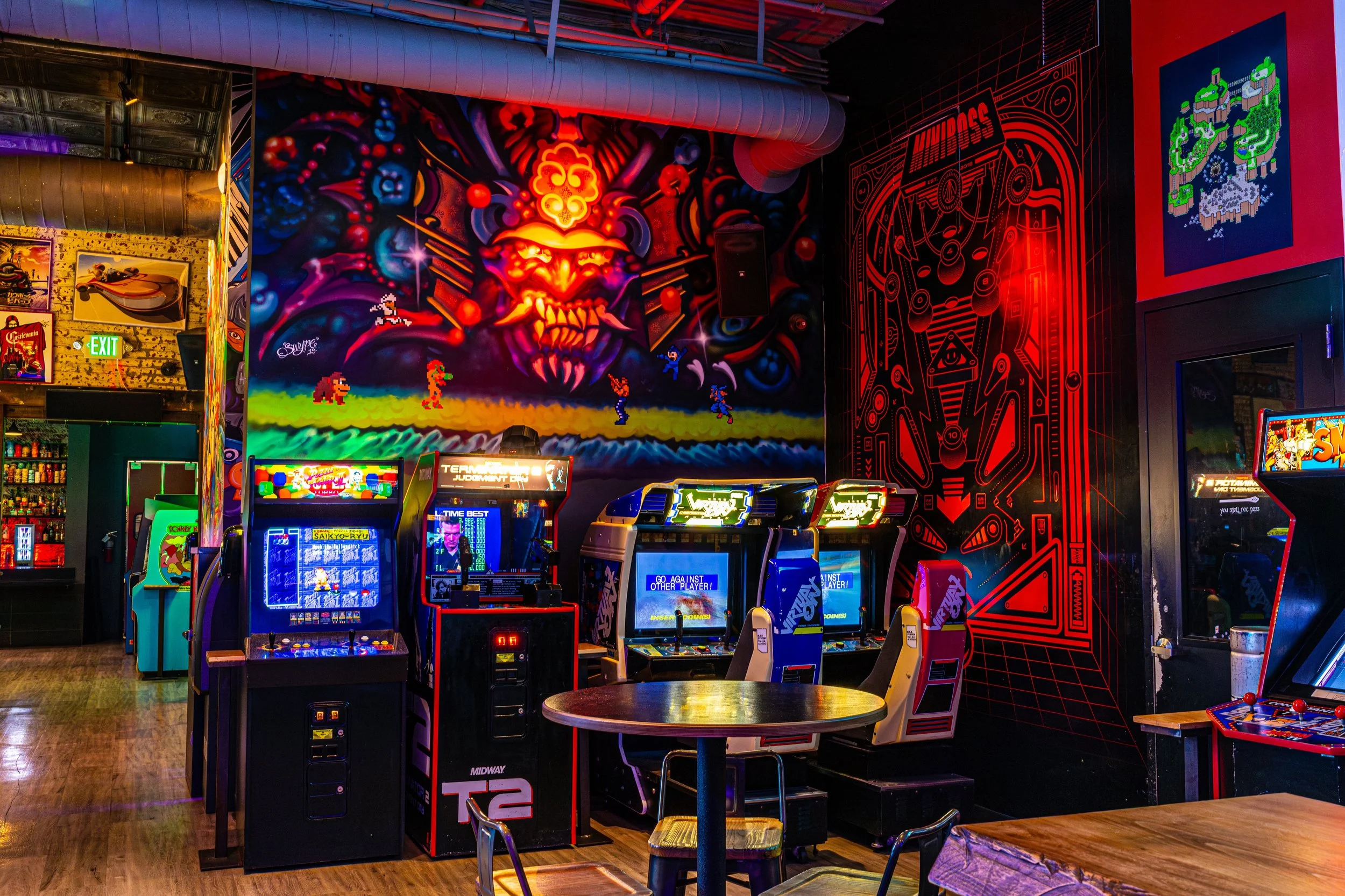 An arcade with multiple video game machines, colorful wall art including pixelated characters from classic video games, and a large vibrant mural of a fiery, demonic face. There are wooden tables and stools nearby.