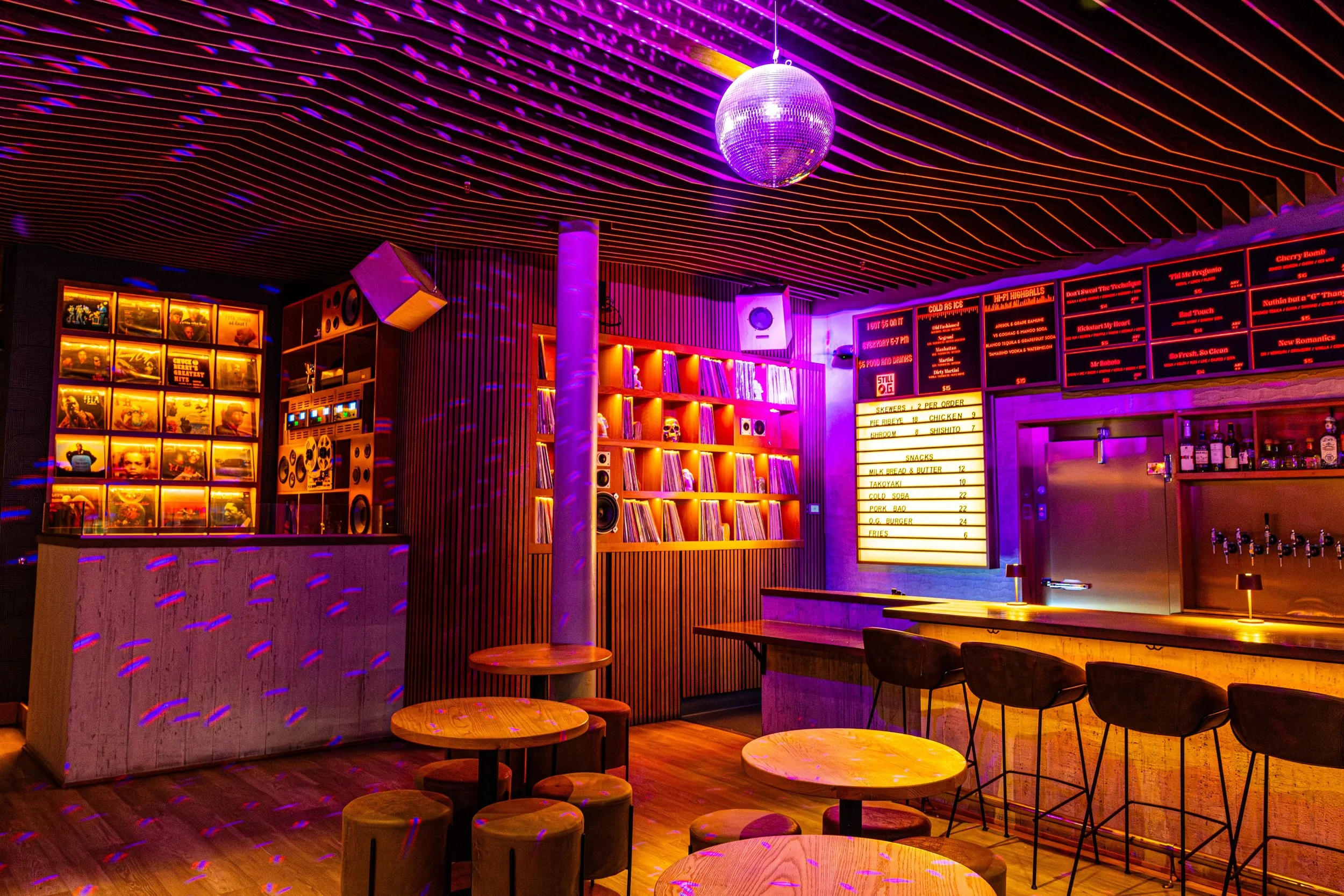 A neon-lit bar or lounge with a spinning disco ball hanging from a ceiling with colorful lighting reflections. There are wooden tables and chairs, and a digital menu board displaying food and drink options in the background.