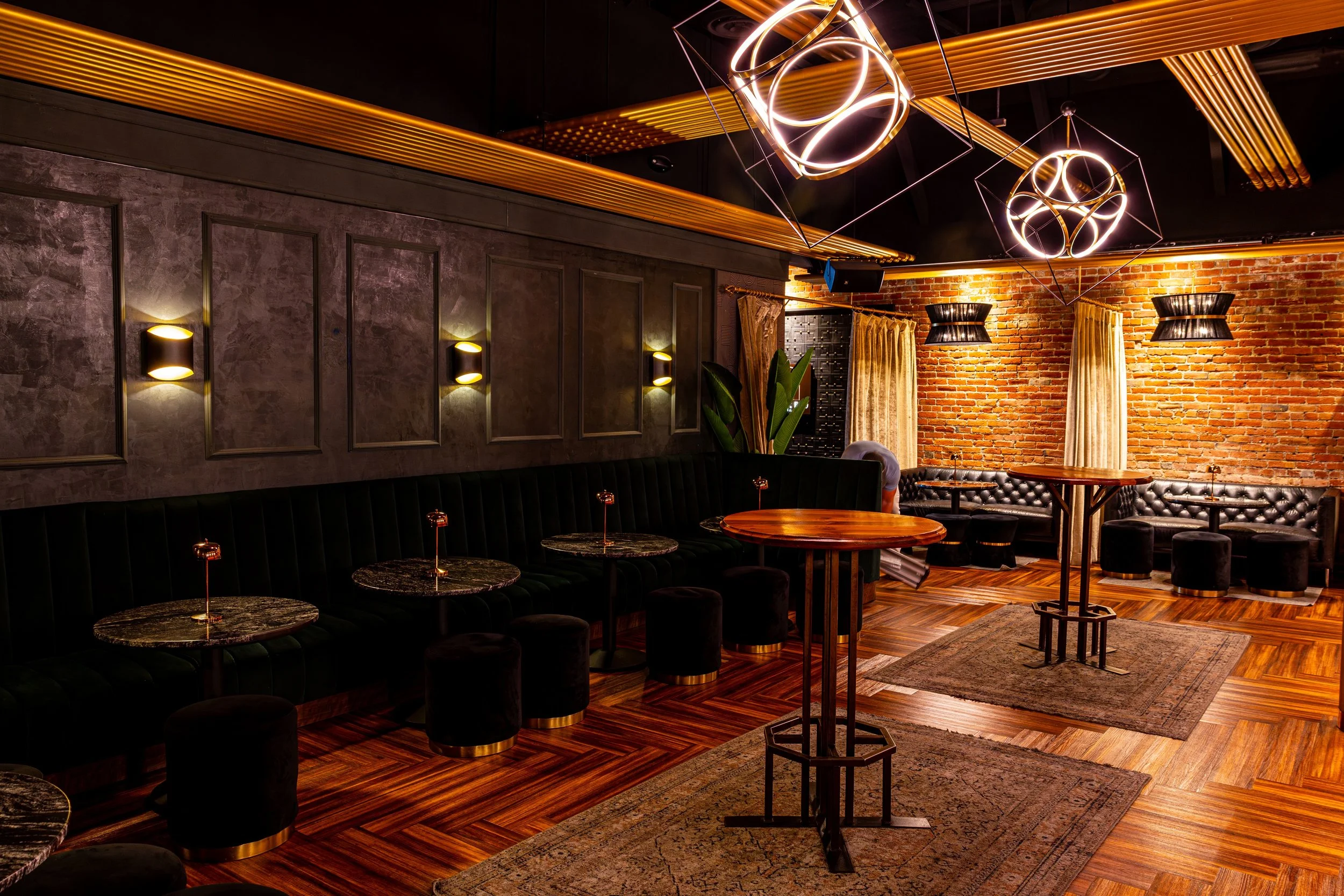 Interior of a stylish lounge or bar with modern geometric light fixtures hanging from the ceiling, warm lighting, exposed brick wall, curtains, and round tables with cushioned seating.